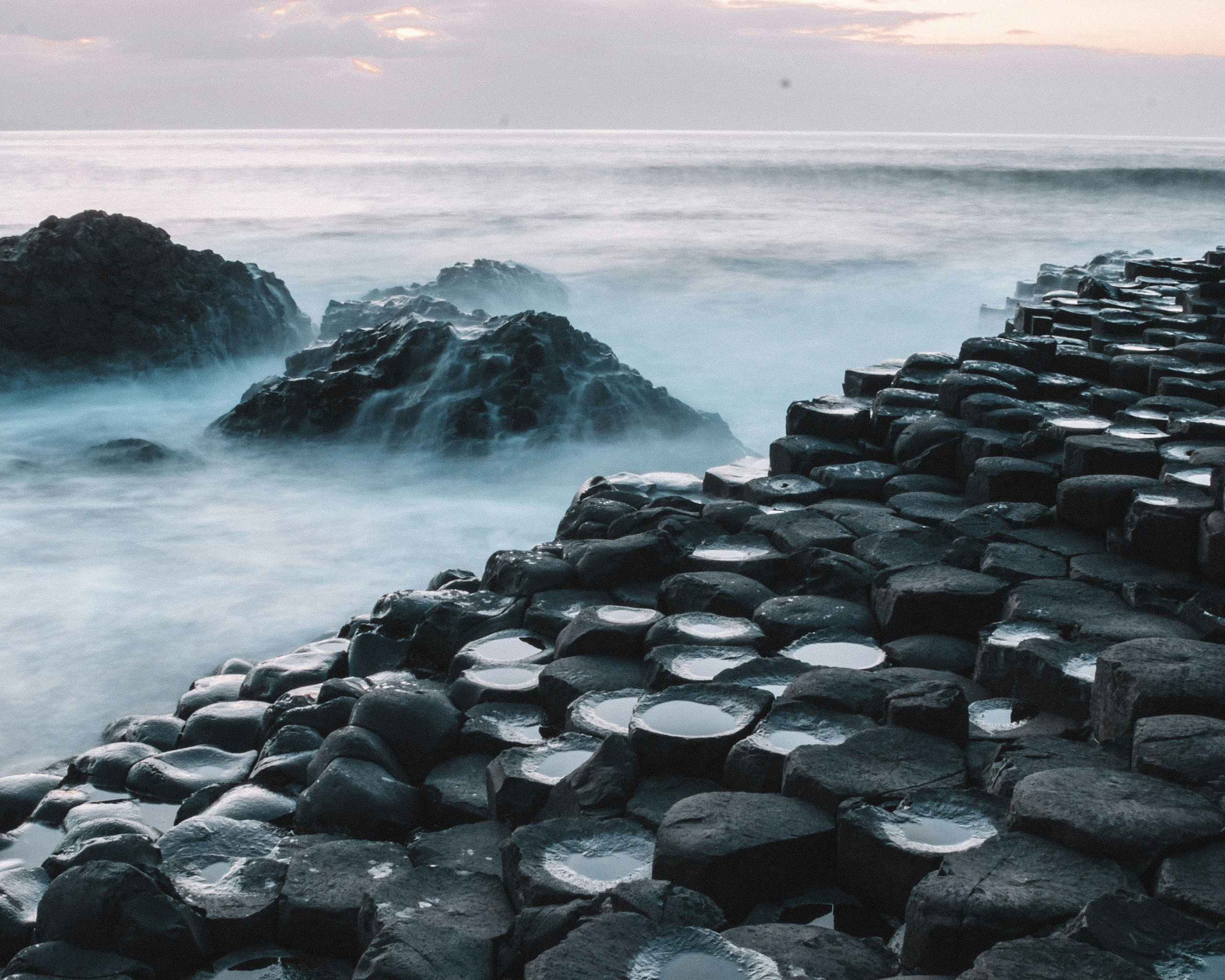 Basaltsäulen des Giant’s Causeway an der Küste von Nordirland, hexagonale Felsformationen am Meer mit brandenden Wellen und mystischer Atmosphäre bei sanftem Abendlicht.