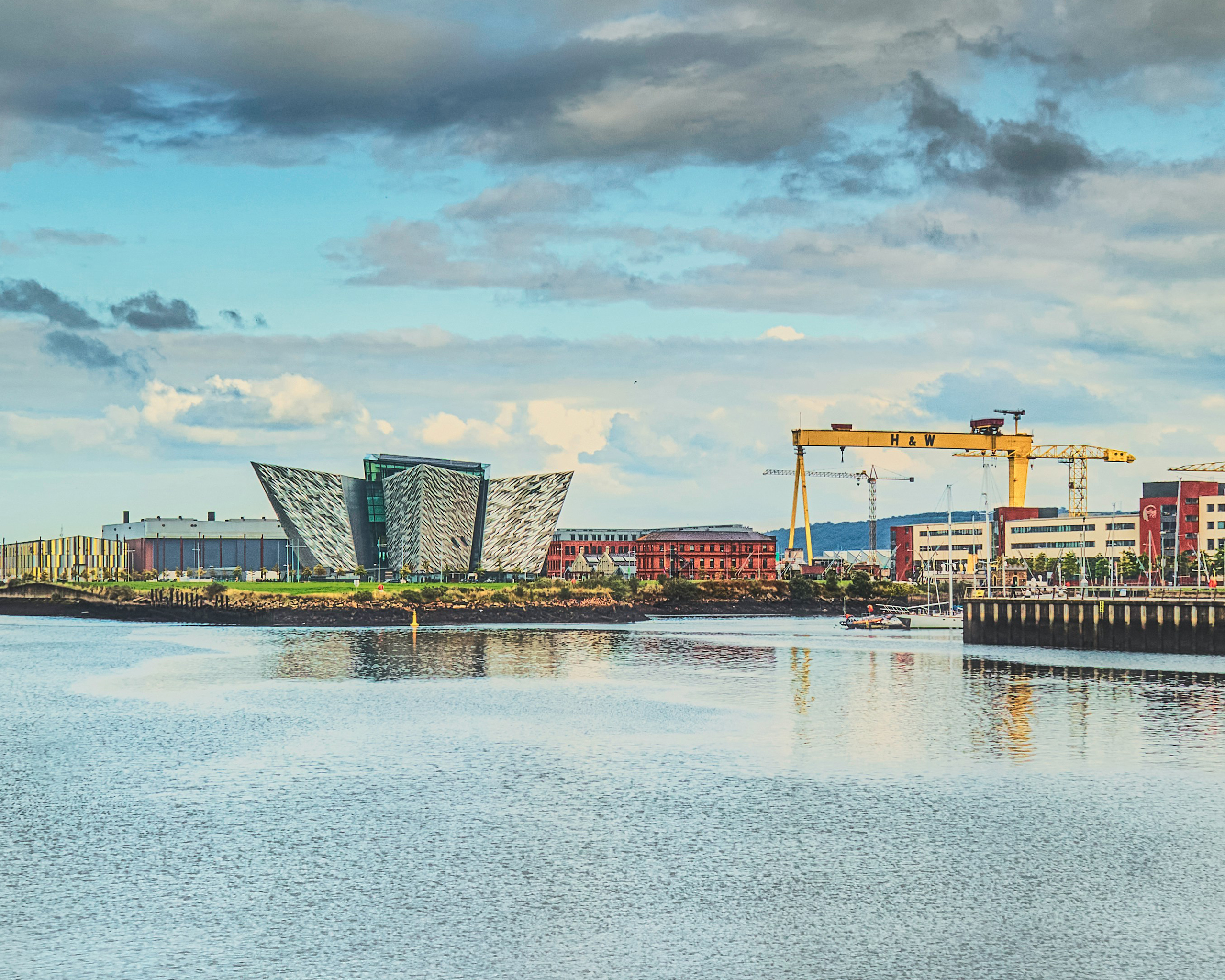Titanic Belfast Museum am Ufer des River Lagan in Belfast, Nordirland, mit markanter moderner Architektur und den gelben Harland-&-Wolff-Kränen im Hintergrund.