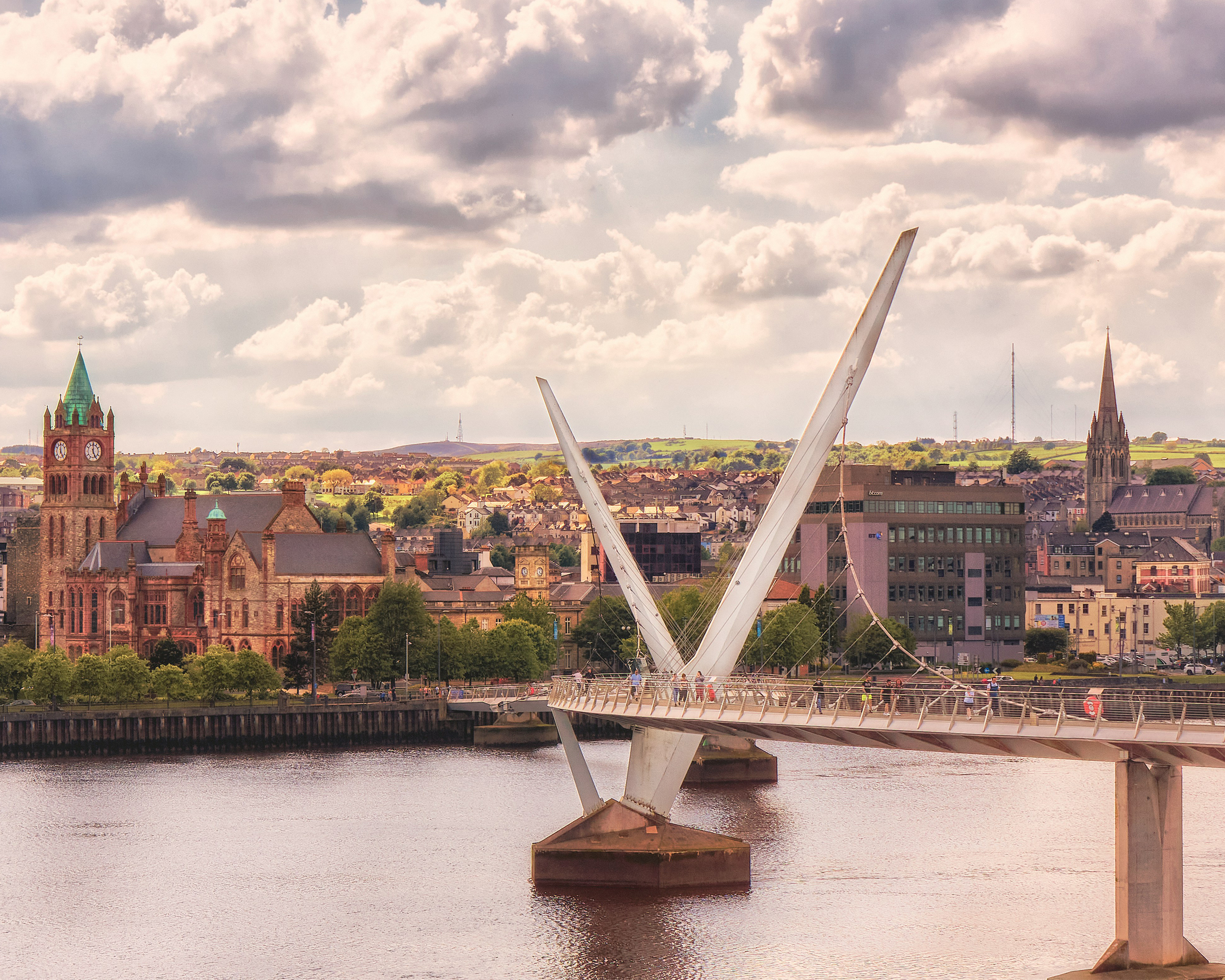Peace Bridge in Derry/Londonderry in Nordirland, moderne Fußgängerbrücke über den River Foyle mit Blick auf die historische Altstadt und markante Kirchengebäude unter bewölktem Himmel.