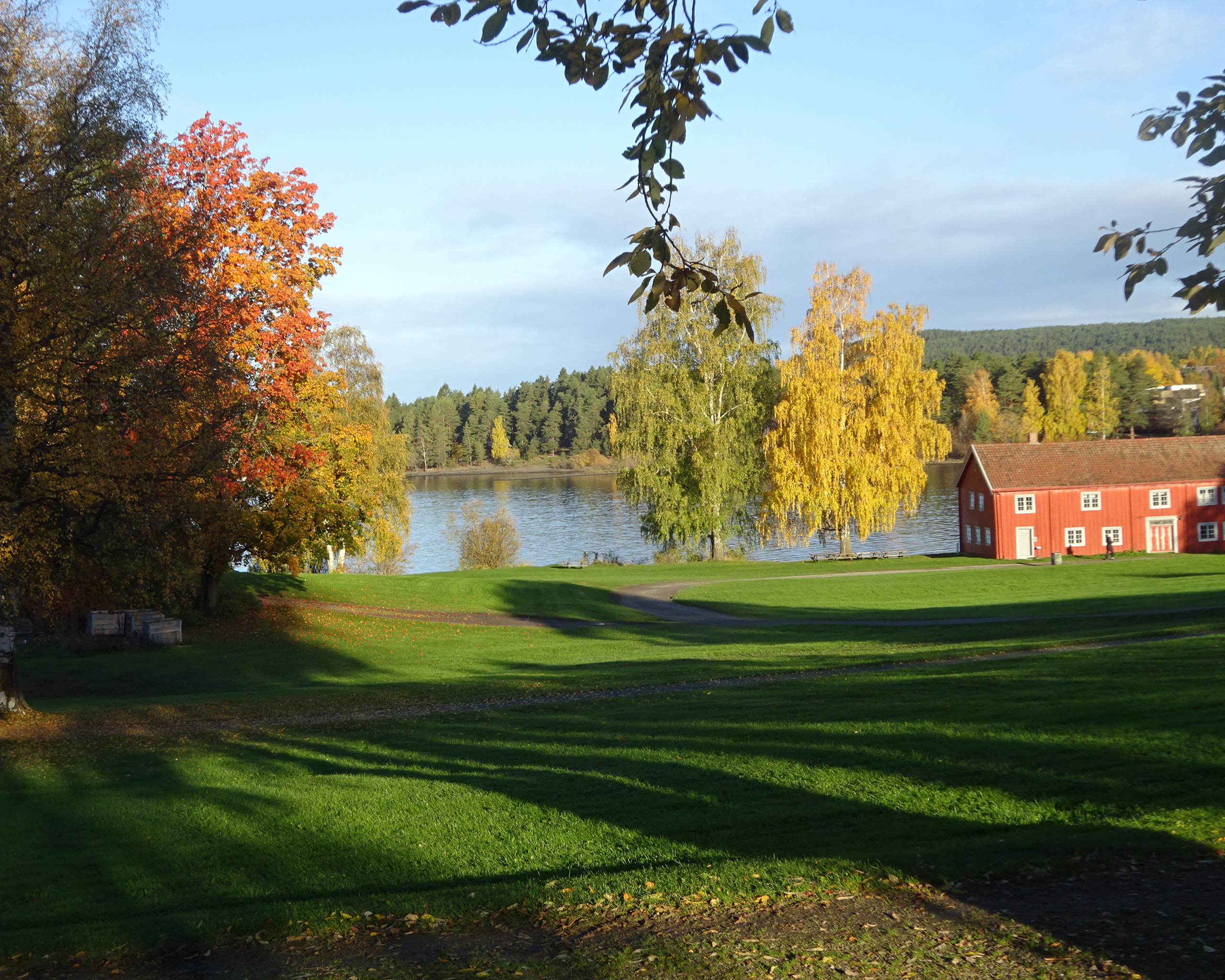 Herbstliche Landschaft am See in Norwegen mit bunten Laubbäumen, grüner Wiese und einem roten Holzhaus am Ufer.