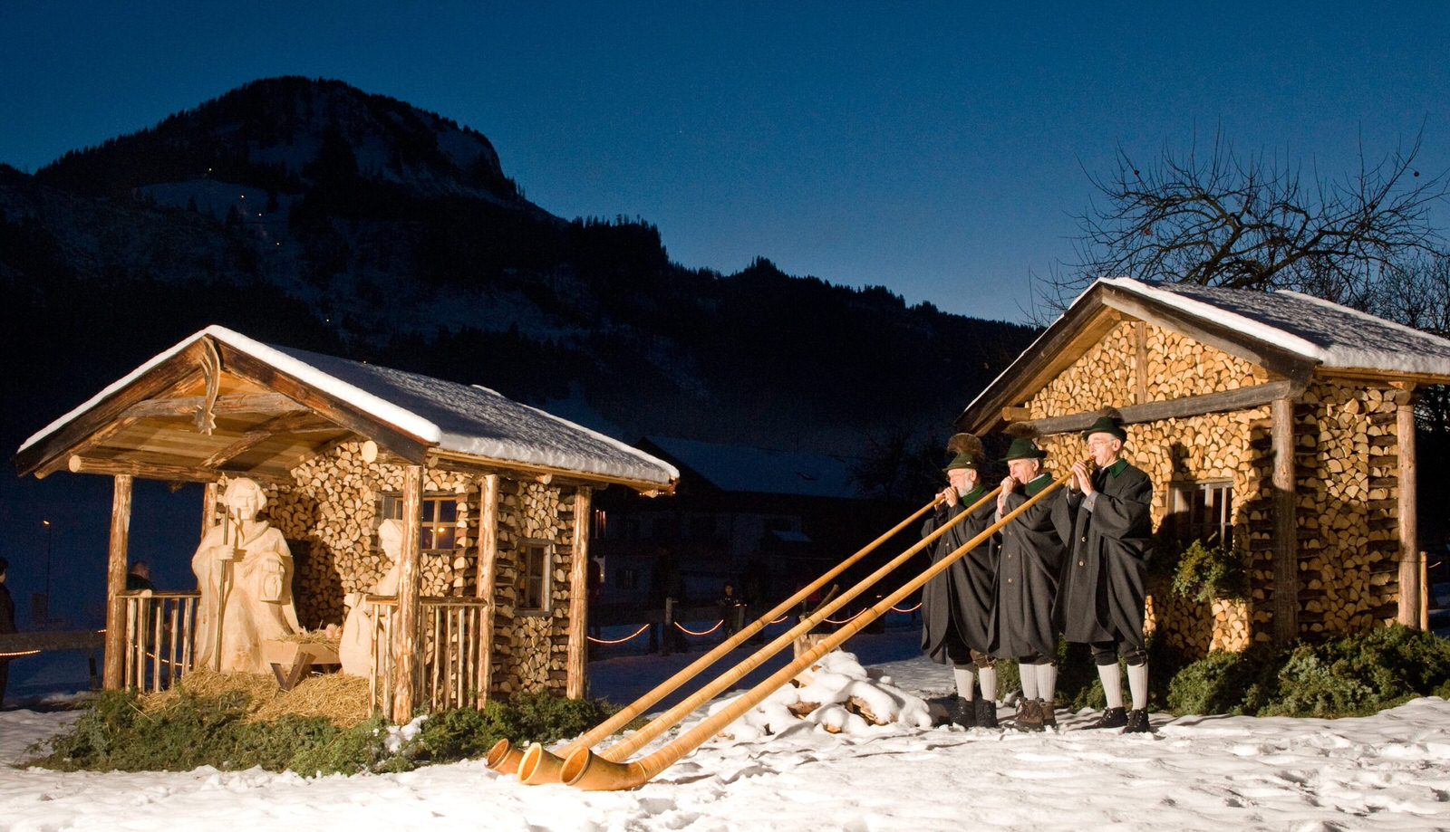 Alphornbläser in traditioneller Tracht spielen vor einer kunstvollen Krippe beim Bad Hindelang Weihnachtsmarkt