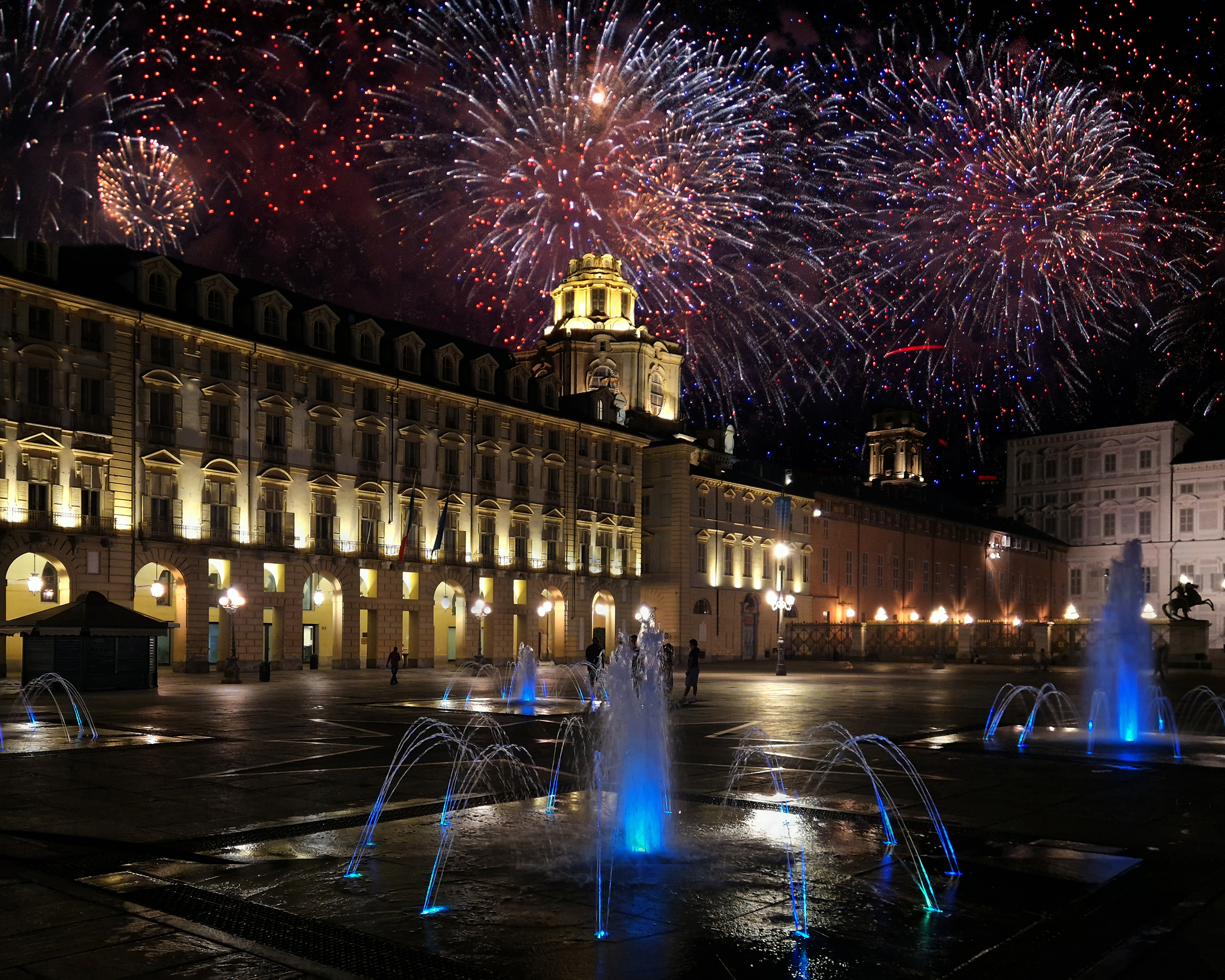 Feuerwerk über der festlich beleuchteten Piazza Castello in Turin während der Silvesternacht mit blau illuminierten Springbrunnen im Vordergrund.