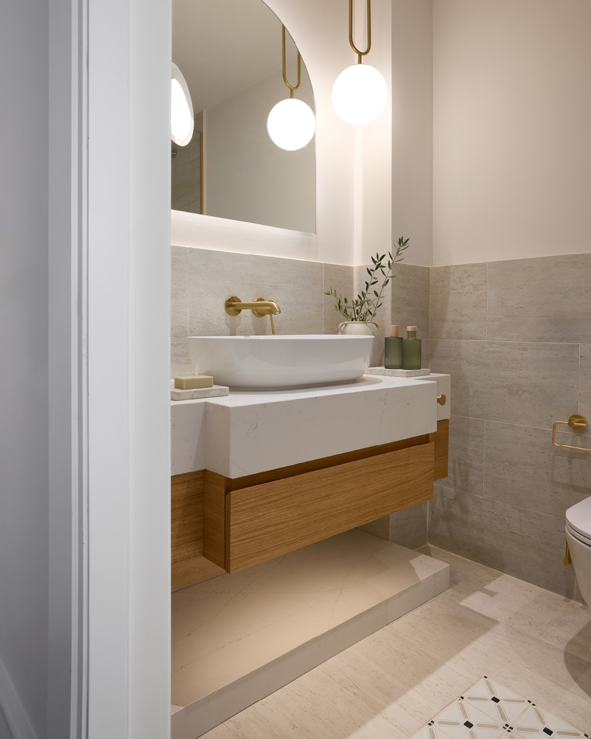 Modernes Hotelbadezimmer mit hellem Waschtisch, rundem Spiegel, goldfarbenen Armaturen und Natursteinfliesen. © Grecotel Filoxenia Kalamata / Hotel