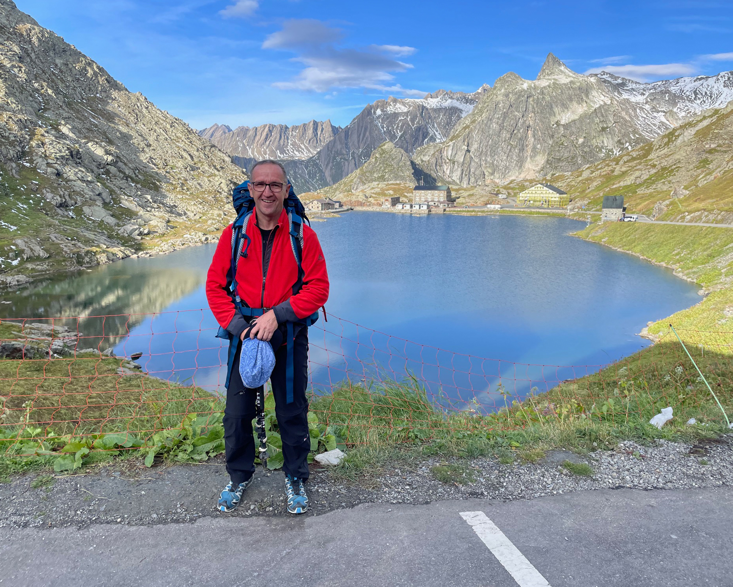 Reiseleiter Harald Pfanner mit Rucksack vor einem Bergsee am Großen Sankt-Bernhard-Pass, umgeben von alpiner Landschaft, Berghütten und schroffen Gipfeln.