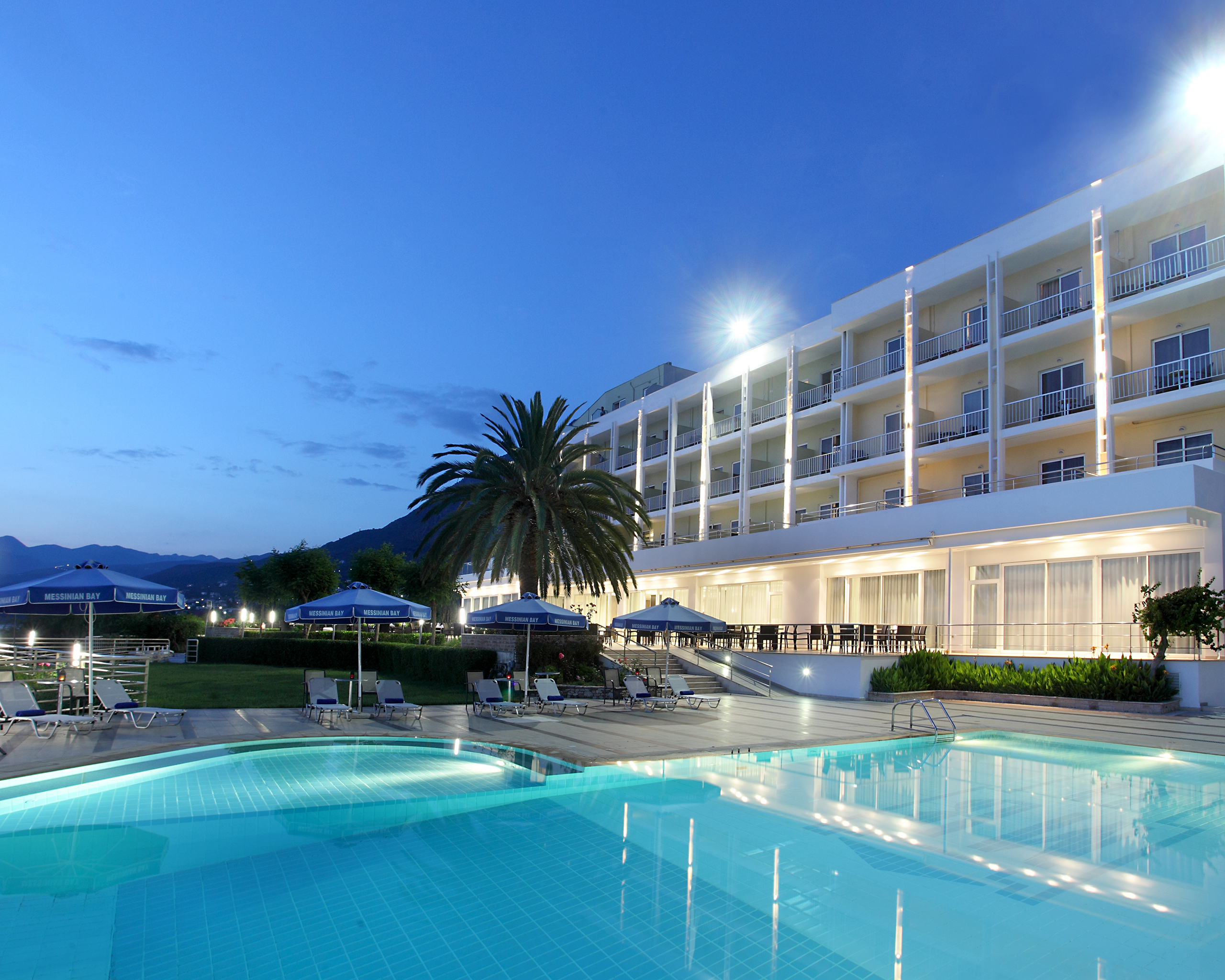 Beleuchtete Hotelanlage des Messinian Bay Hotels in Kalamata mit Pool, Palmen und moderner Architektur in der Abenddämmerung. © Idealtours