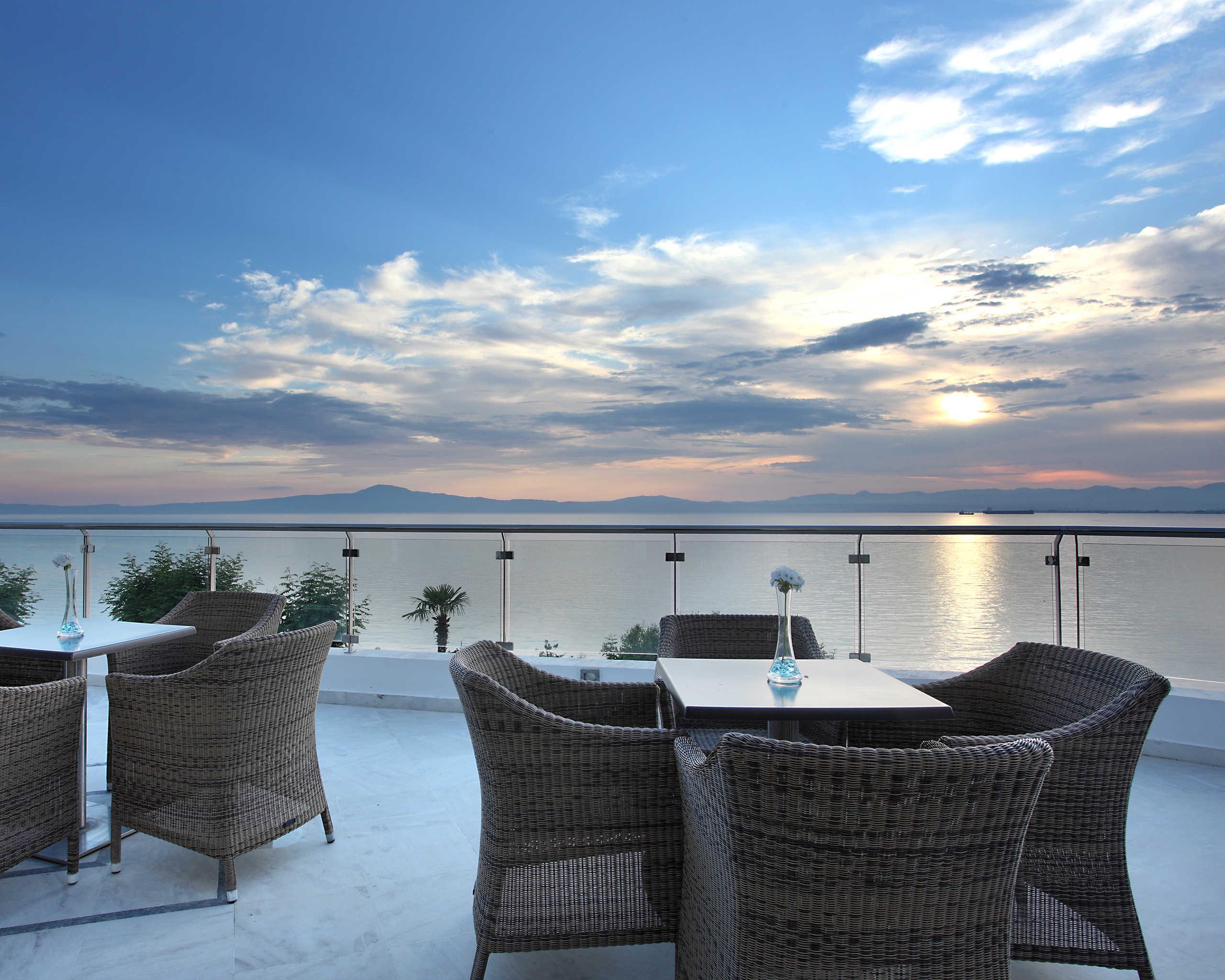 Hotelterrasse mit Sitzgelegenheiten und Glasgeländer mit Blick auf das Meer bei Sonnenuntergang, Messinian Bay Hotel, Kalamata. © Idealtours