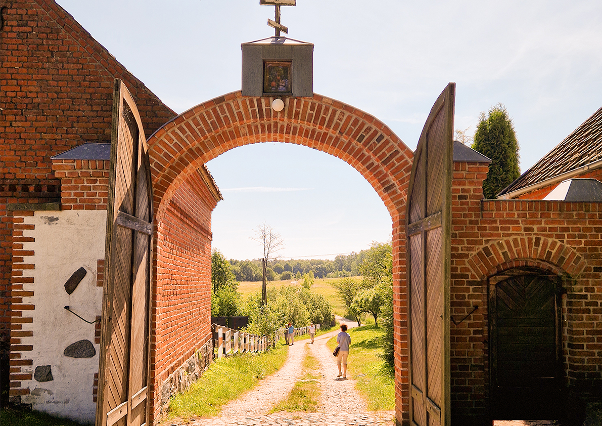Historisches Backsteintor mit geöffneten Holztoren und Blick auf einen Feldweg in der polnischen Landschaft.