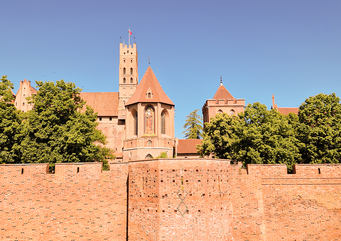 Mächtige Ordensburg aus rotem Backstein hinter einer Wehrmauer, umgeben von grünen Bäumen in Polen.