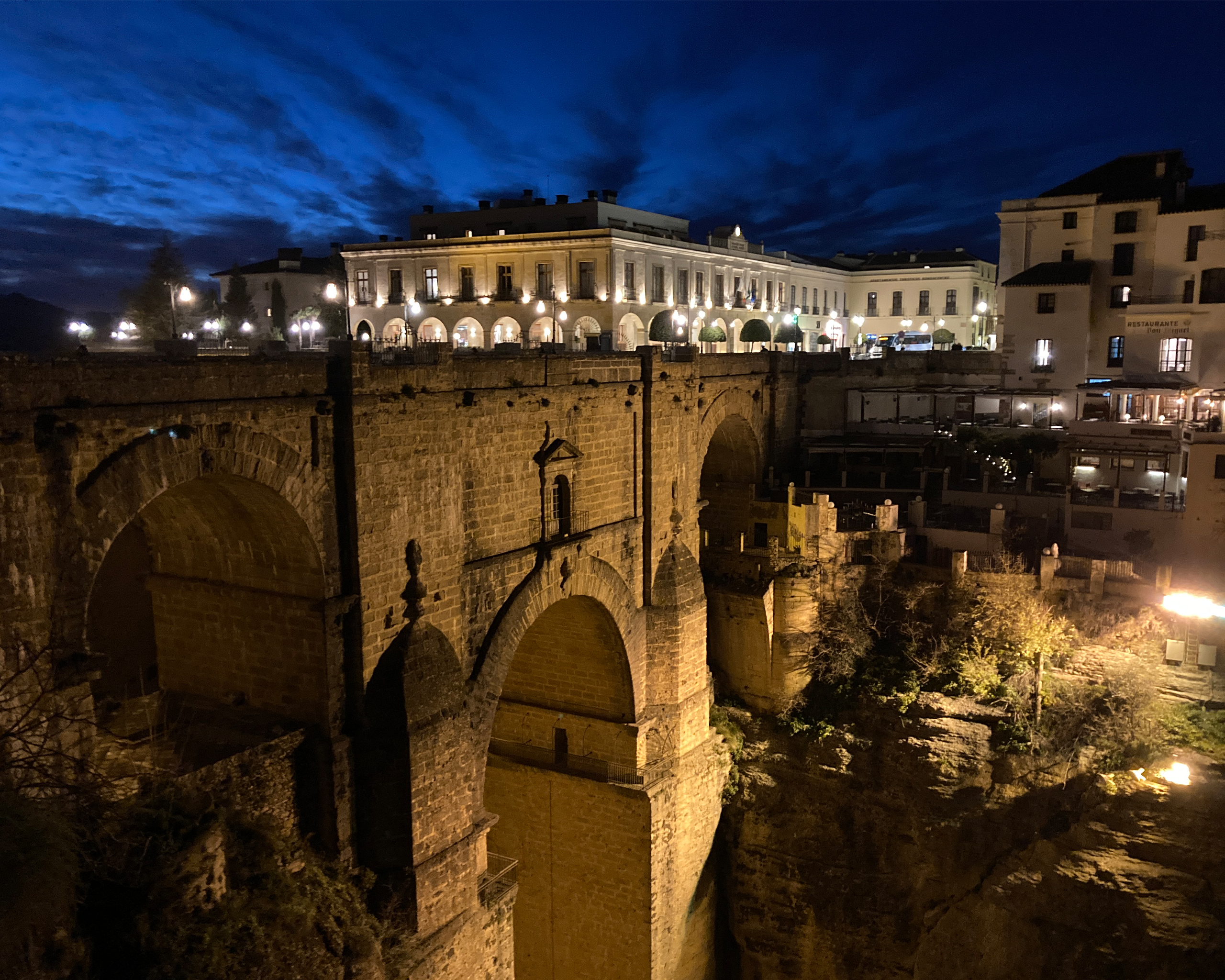 Beleuchtete historische Steinbrücke über eine tiefe Schlucht bei Nacht, dahinter erleuchtete Altstadt mit Arkaden und Gebäuden unter dramatischem Abendhimmel in Andalusien.