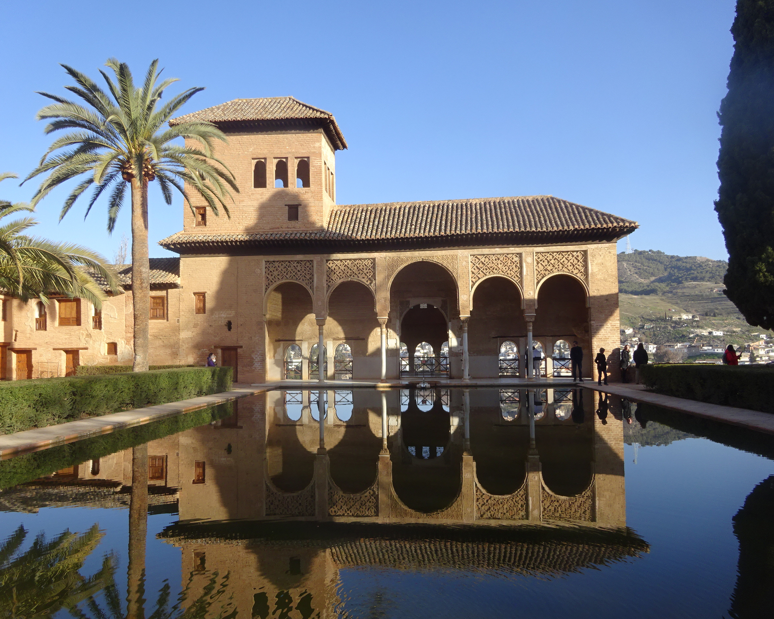 Maurischer Innenhof der Alhambra in Granada mit kunstvollen Arkaden, Spiegelbecken und Palmen, symmetrisch gespiegelt im ruhigen Wasser unter blauem Himmel.