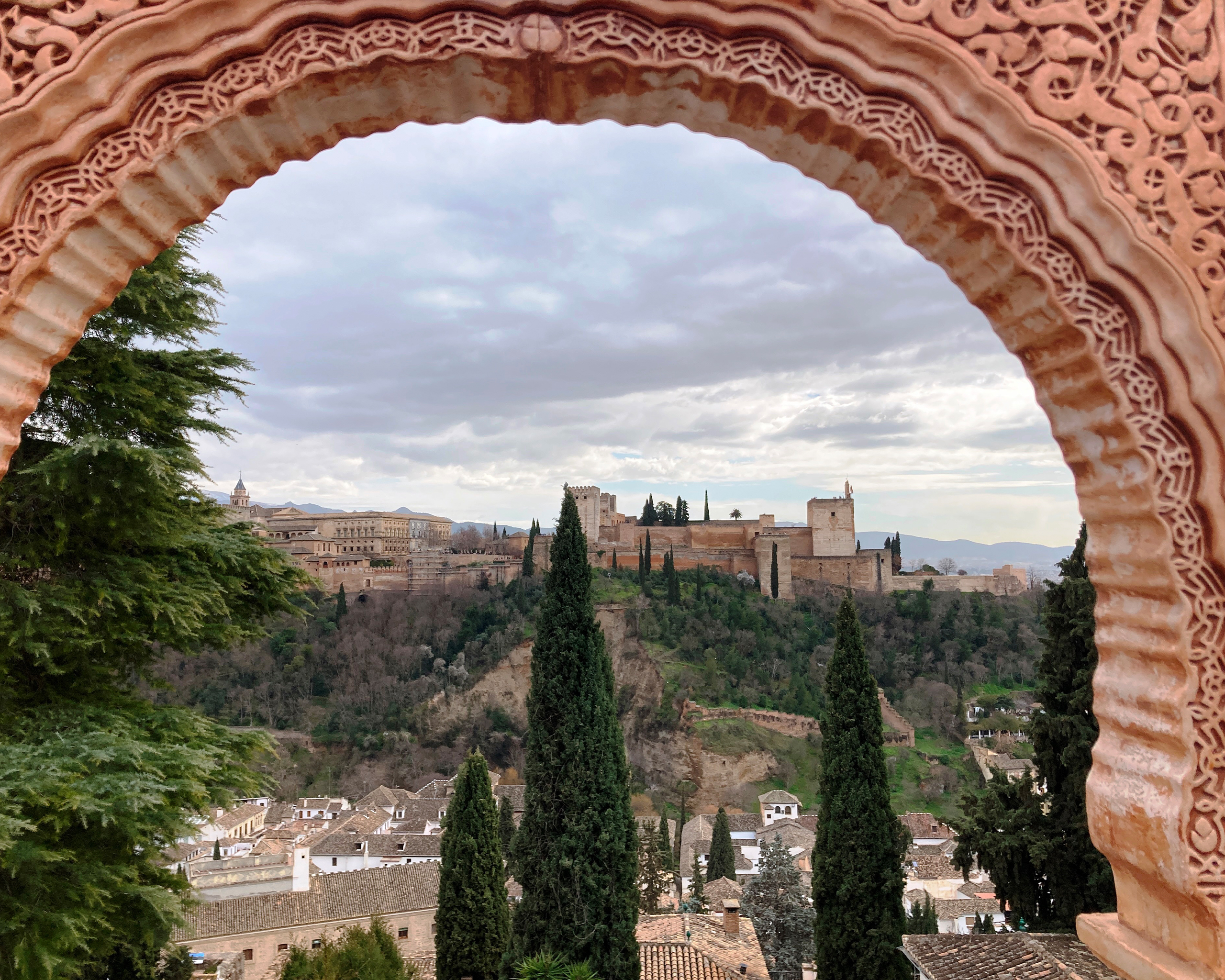 Blick durch einen kunstvoll verzierten maurischen Torbogen auf die Alhambra von Granada, umgeben von Zypressen, Hügeln und der historischen Altstadt.