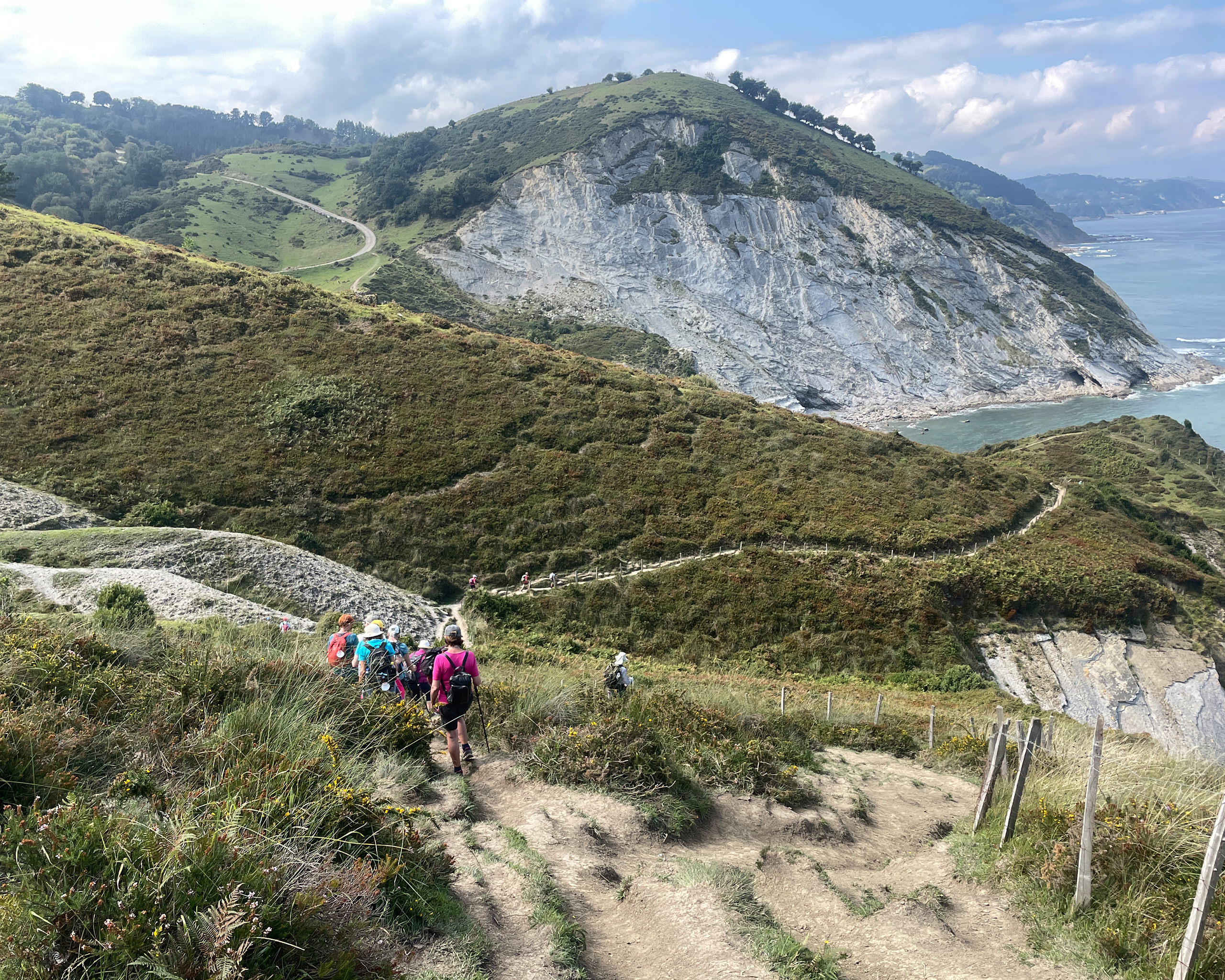 Pilger steigen auf einem schmalen Pfad durch hügelige Landschaft mit steilen Klippen und Blick auf den Atlantik entlang des Camino del Norte in Spanien.