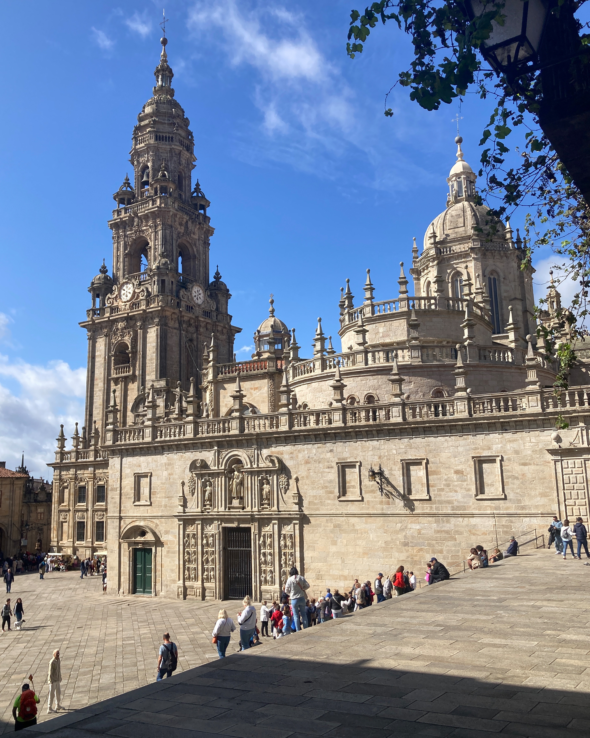 Imposante Kathedrale von Santiago de Compostela mit reich verzierter Fassade und Glockenturm, Pilger und Besucher auf dem weiten Vorplatz unter blauem Himmel.