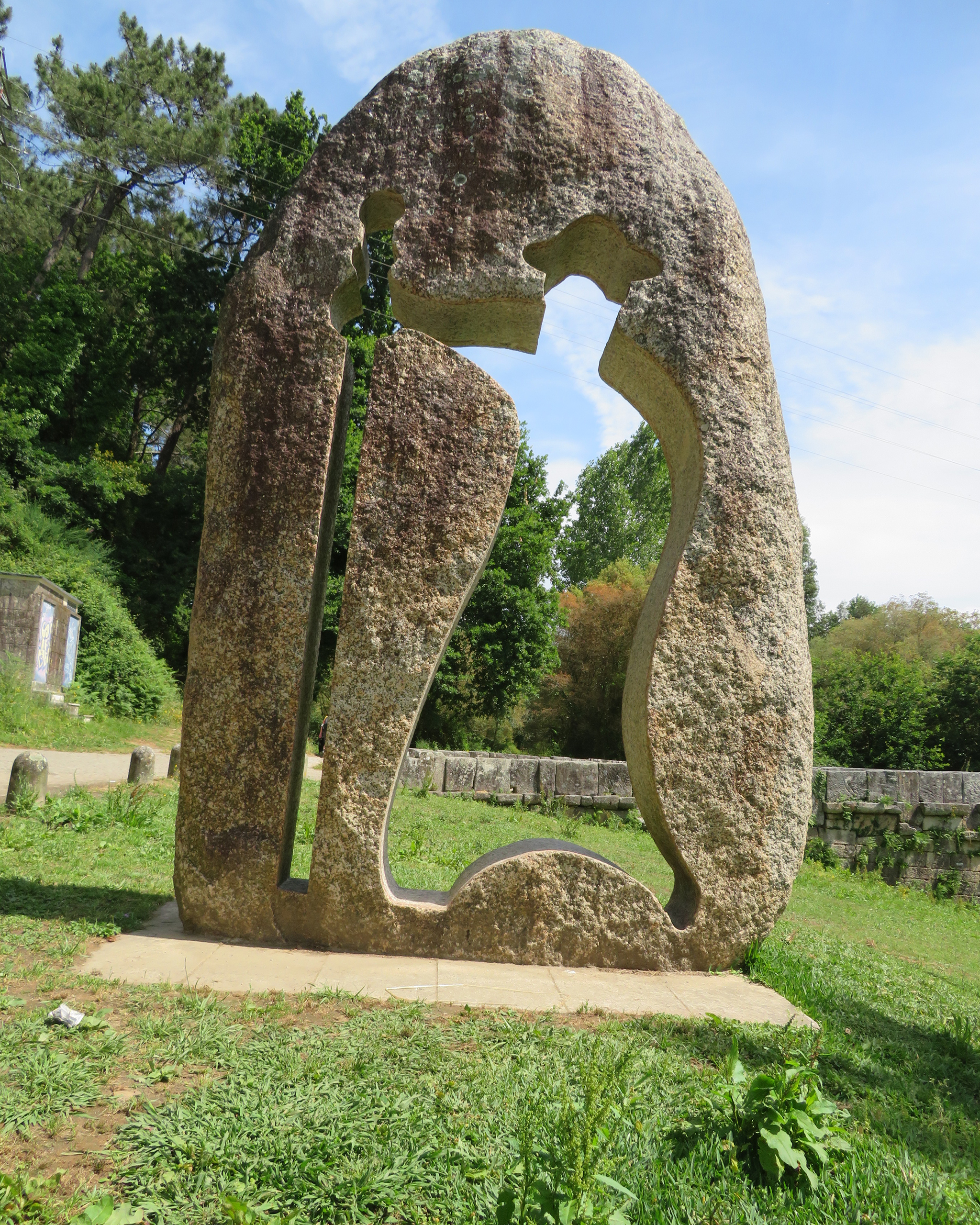 Große abstrakte Steinskulptur mit Durchbruch in Form eines stilisierten Pilgers, aufgestellt in einer grünen Parklandschaft entlang des Jakobswegs bei Giengen an der Brenz.