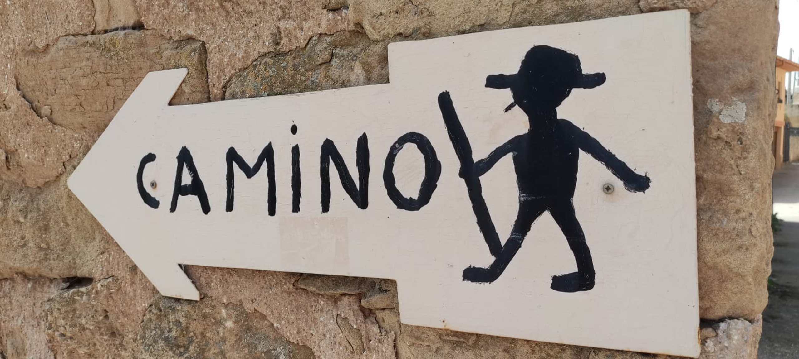 Blick auf ein handgemaltes weißes Wegschild mit Pfeil und Pilgerfigur, beschriftet mit dem Wort „Camino“, als Markierung des Jakobswegs an einer Steinmauer.