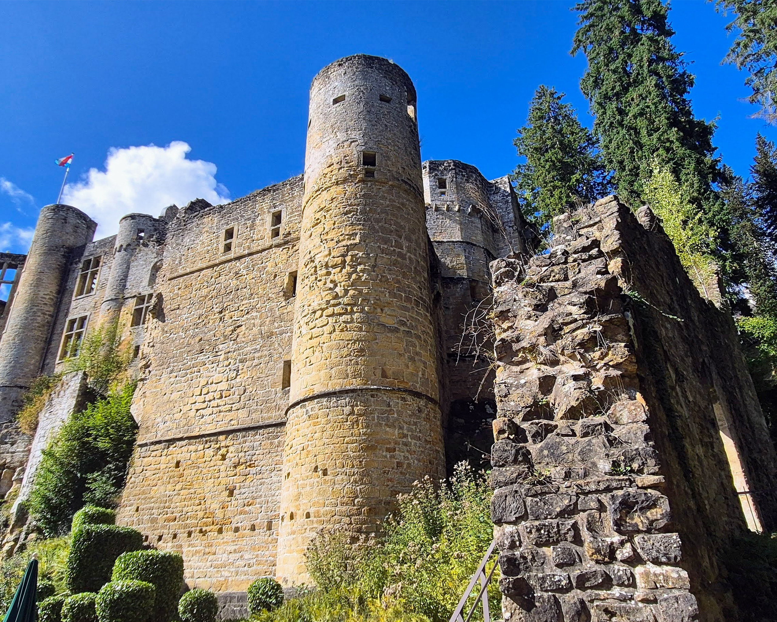 Mittelalterliche Burgruine mit runden Wehrtürmen aus hellem Stein, umgeben von Bäumen unter strahlend blauem Himmel in Luxemburg.