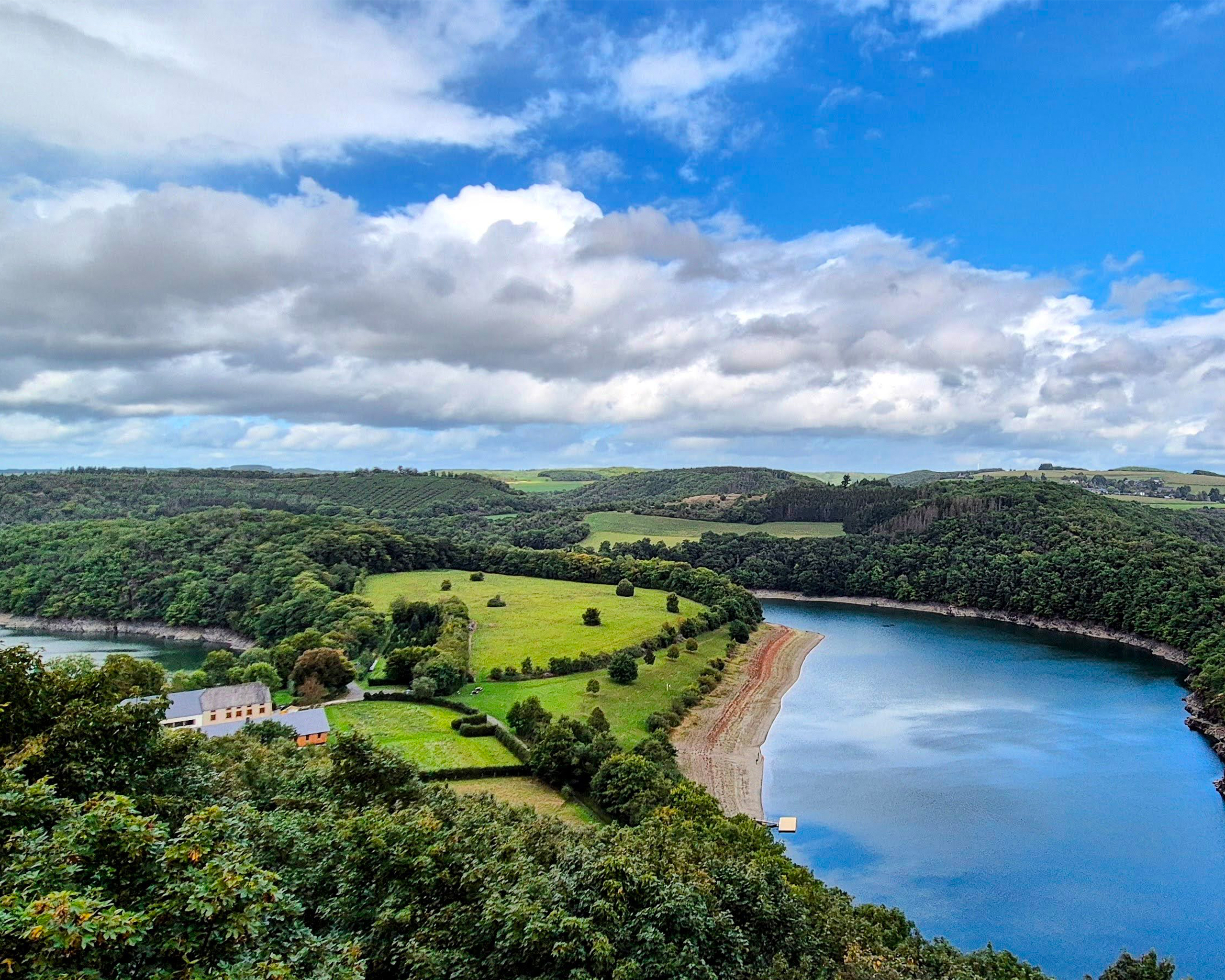 Weite Seenlandschaft mit geschwungenem Ufer, grünen Wiesen und bewaldeten Hügeln im Naturpark Obersauer in Luxemburg.
