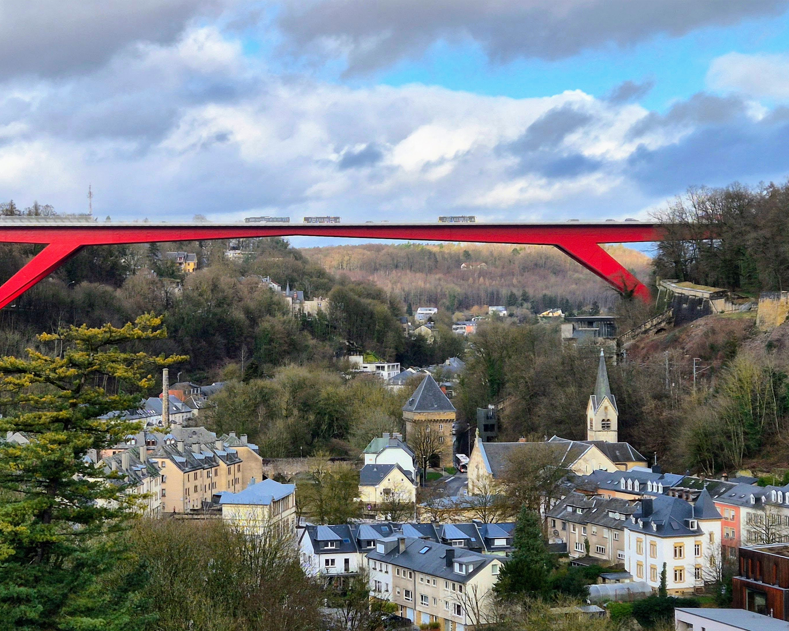 Panoramablick auf eine luxemburgische Stadt mit Kirche und Wohnhäusern, darüber eine markante rote Brücke, eingebettet in ein bewaldetes Tal.