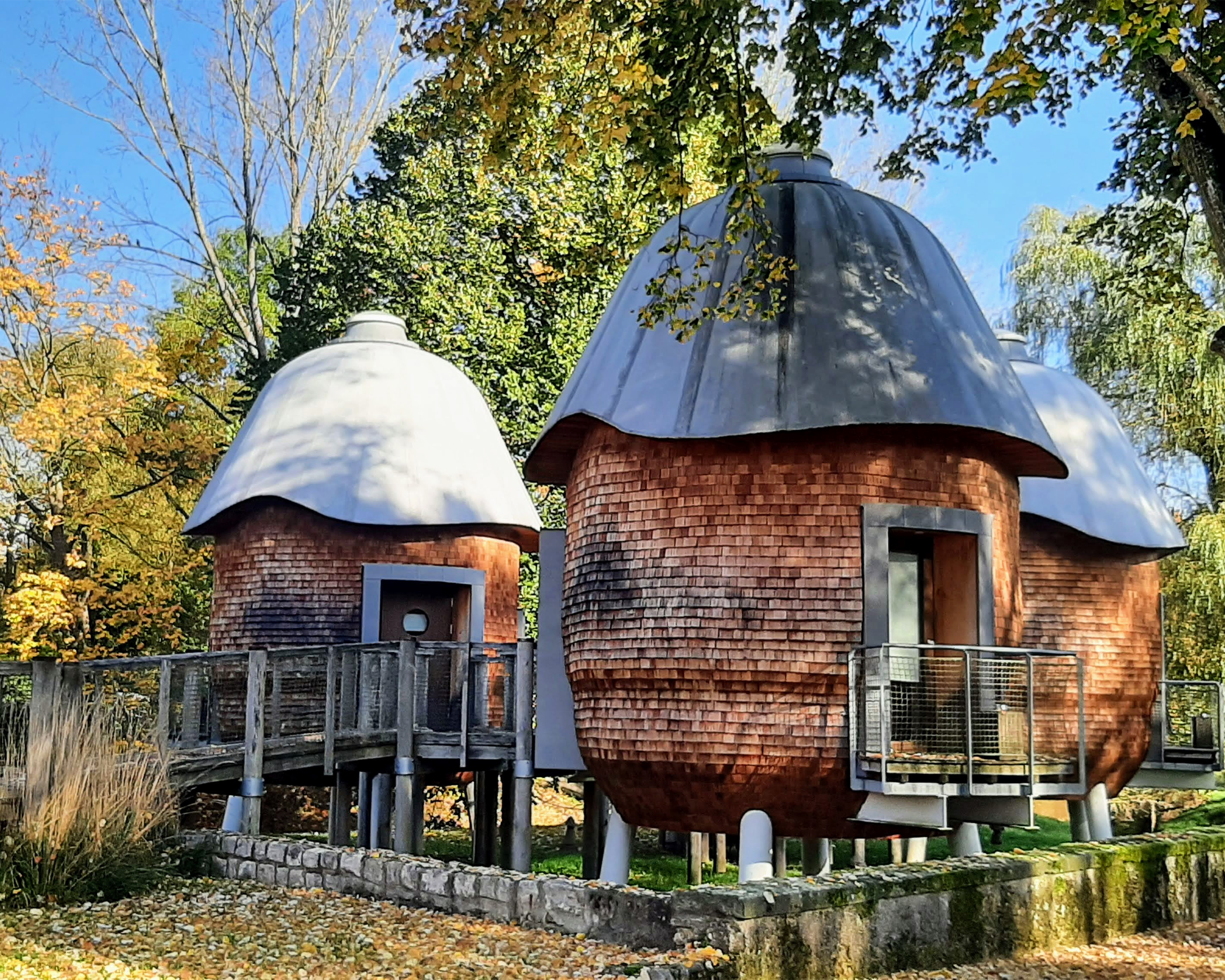 Moderne runde Holzbungalows auf Stelzen mit Metallkuppeldächern, idyllisch gelegen inmitten eines herbstlichen Parks in Luxemburg.