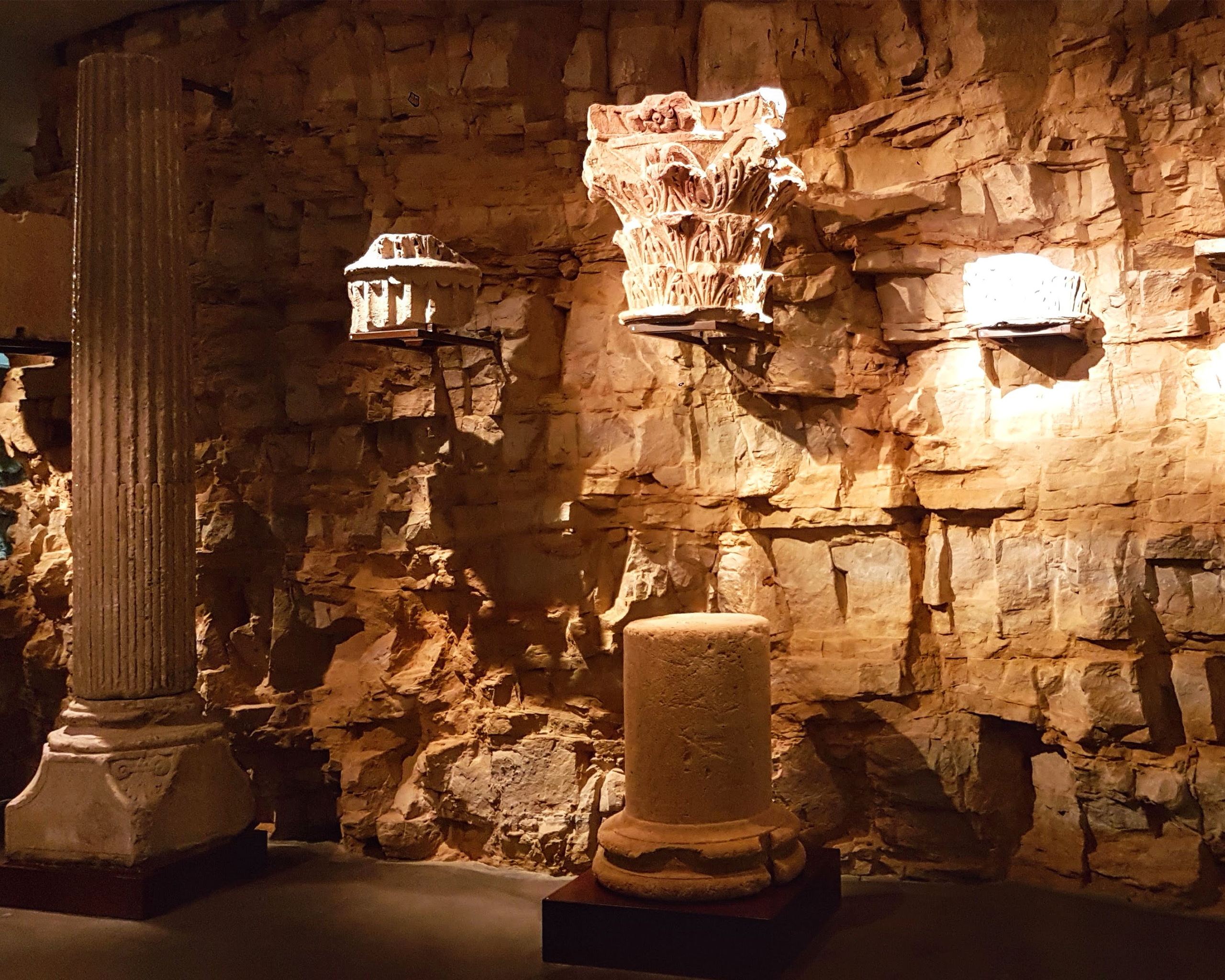 Beleuchteter Museumsraum mit antiken Steinsäulen und kunstvoll verzierten Kapitellen vor einer historischen Felswand in Luxemburg.