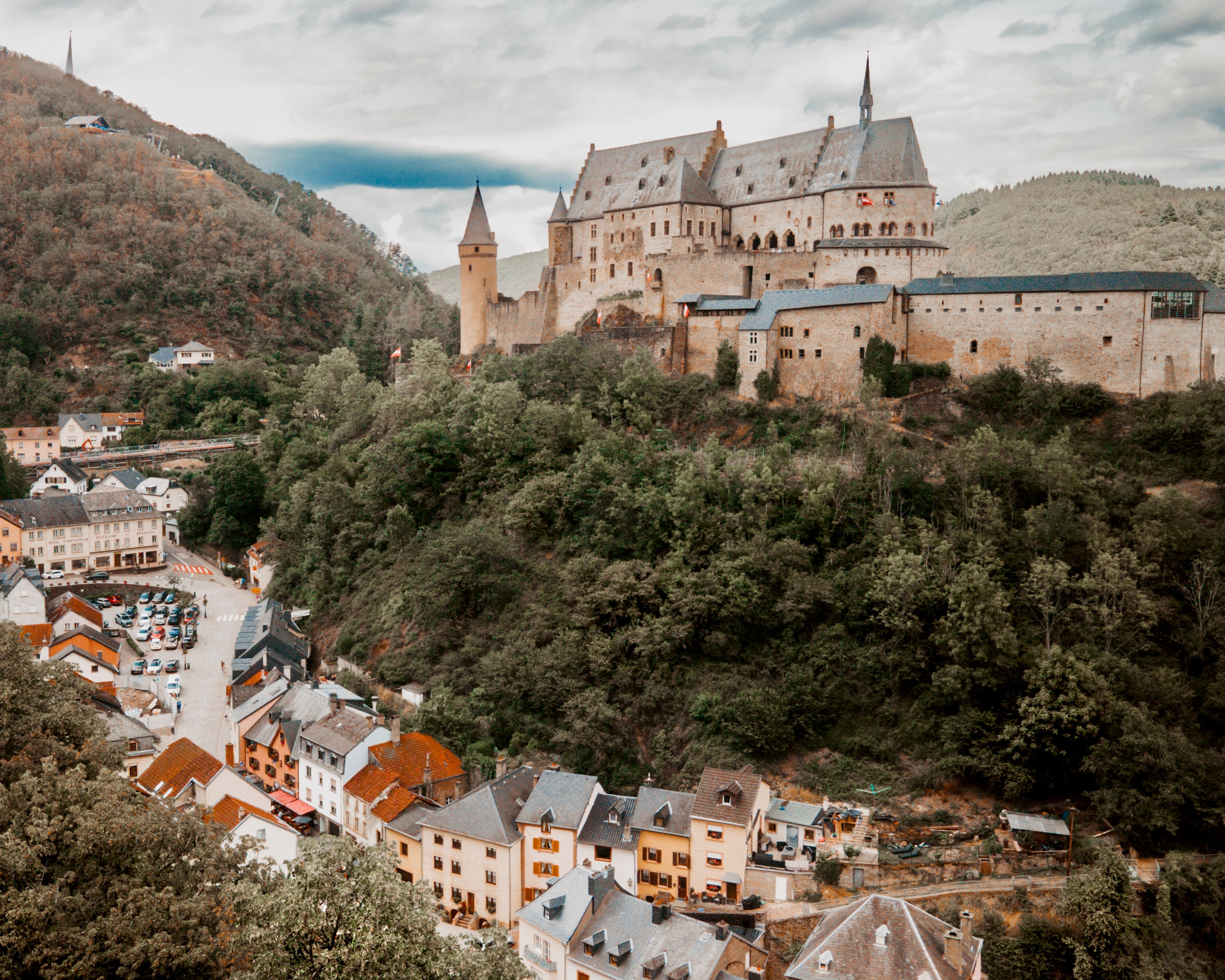 Malerischer Blick auf eine imposante Burganlage hoch über einer Stadt, umgeben von bewaldeten Hügeln im Großherzogtum Luxemburg.