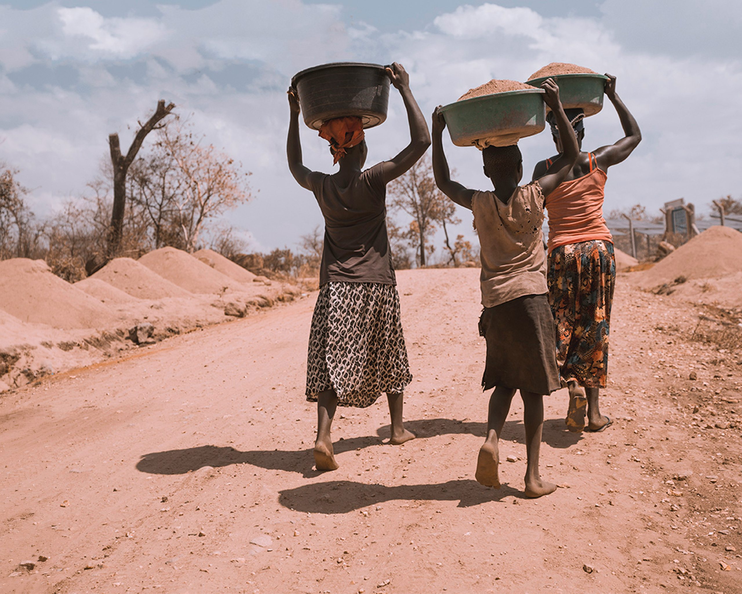 Drei Frauen tragen große Körbe auf dem Kopf entlang einer staubigen Straße in einer ländlichen Region Ugandas.