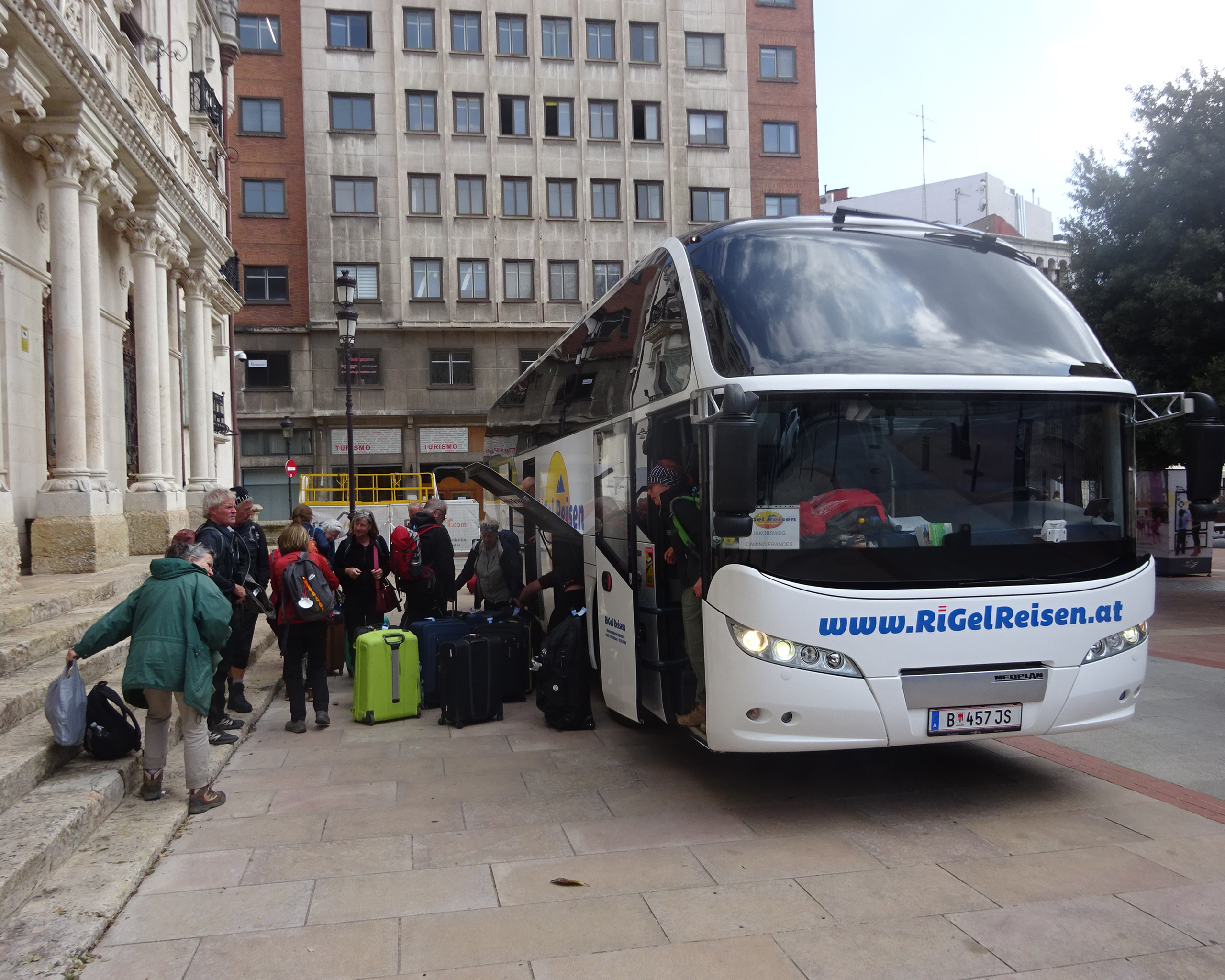 Pilgergruppe beim Einladen des Gepäcks in einen modernen Reisebus von RiGel Reisen in einer spanischen Stadt, sichtbar der Schriftzug „www.RiGelReisen.at“ auf der Busfront, Auftakt einer Camino Francés Pilgerreise.