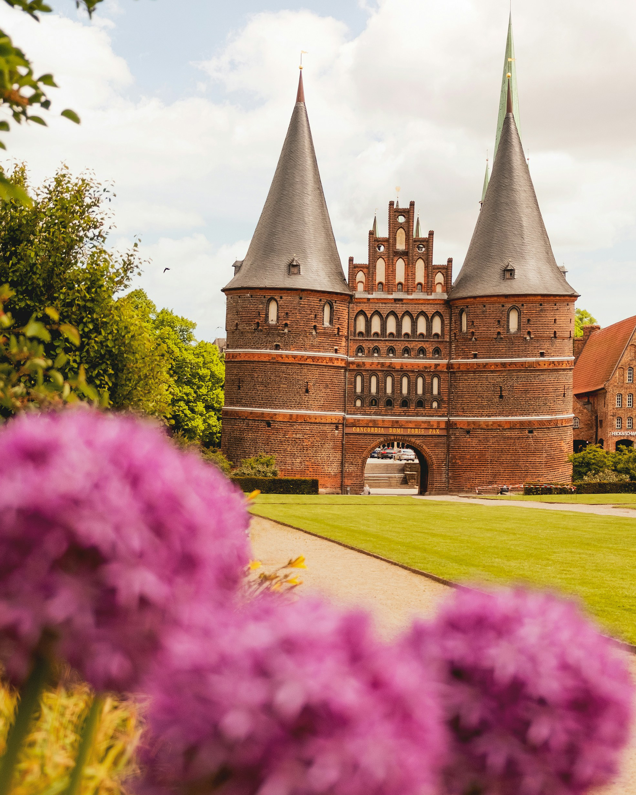 Historisches Holstentor in Lübeck mit zwei markanten Rundtürmen, eingerahmt von grüner Parkanlage und blühenden Blumen im Vordergrund.