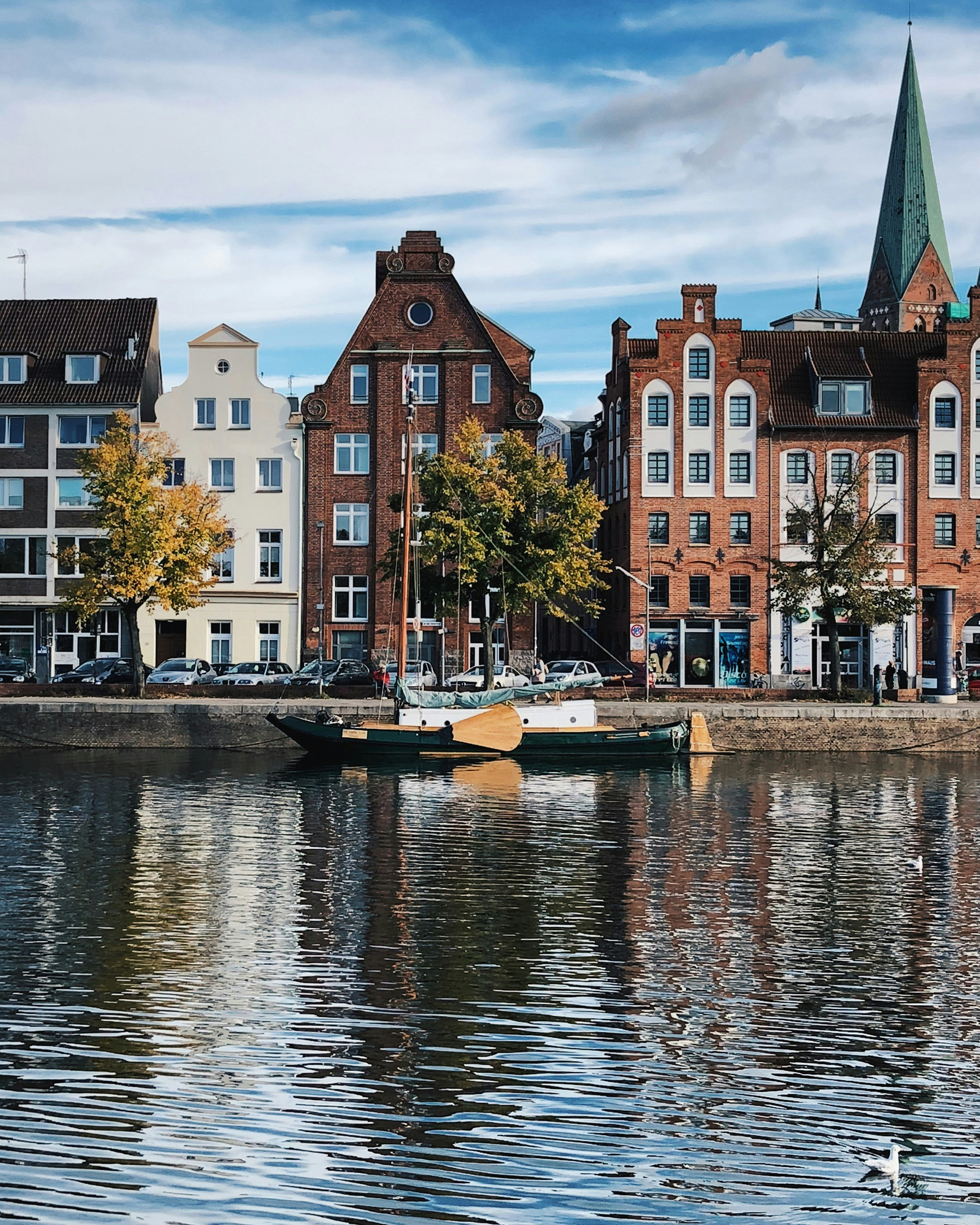 Traditionelle Backsteinhäuser an der Trave in Lübeck, mit kleinem Boot im Vordergrund und Spiegelungen im ruhigen Wasser.