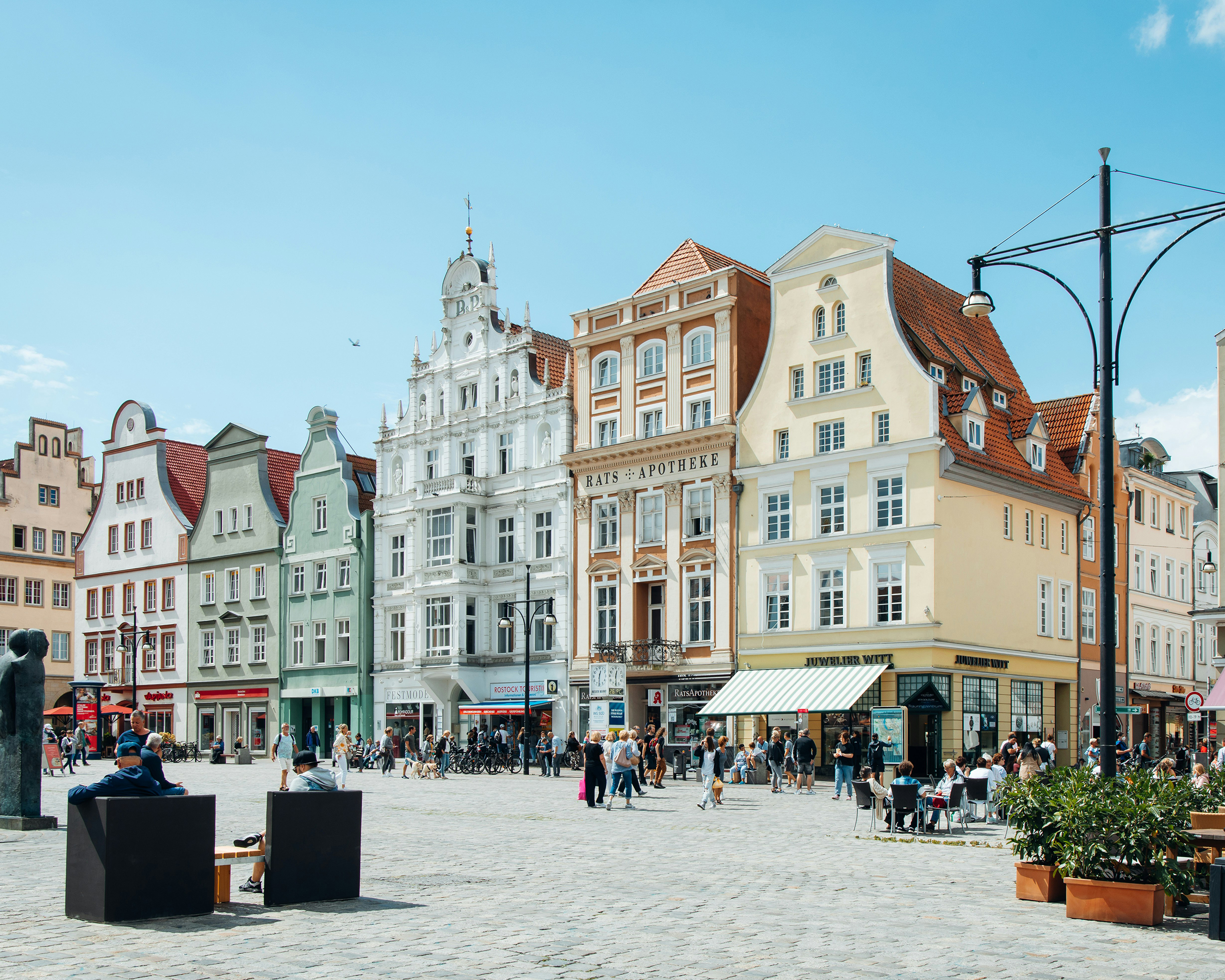 Bunte historische Giebelhäuser am Neuen Markt in Rostock, mit belebtem Platz, Straßencafés und Spaziergängern bei sonnigem Wetter.