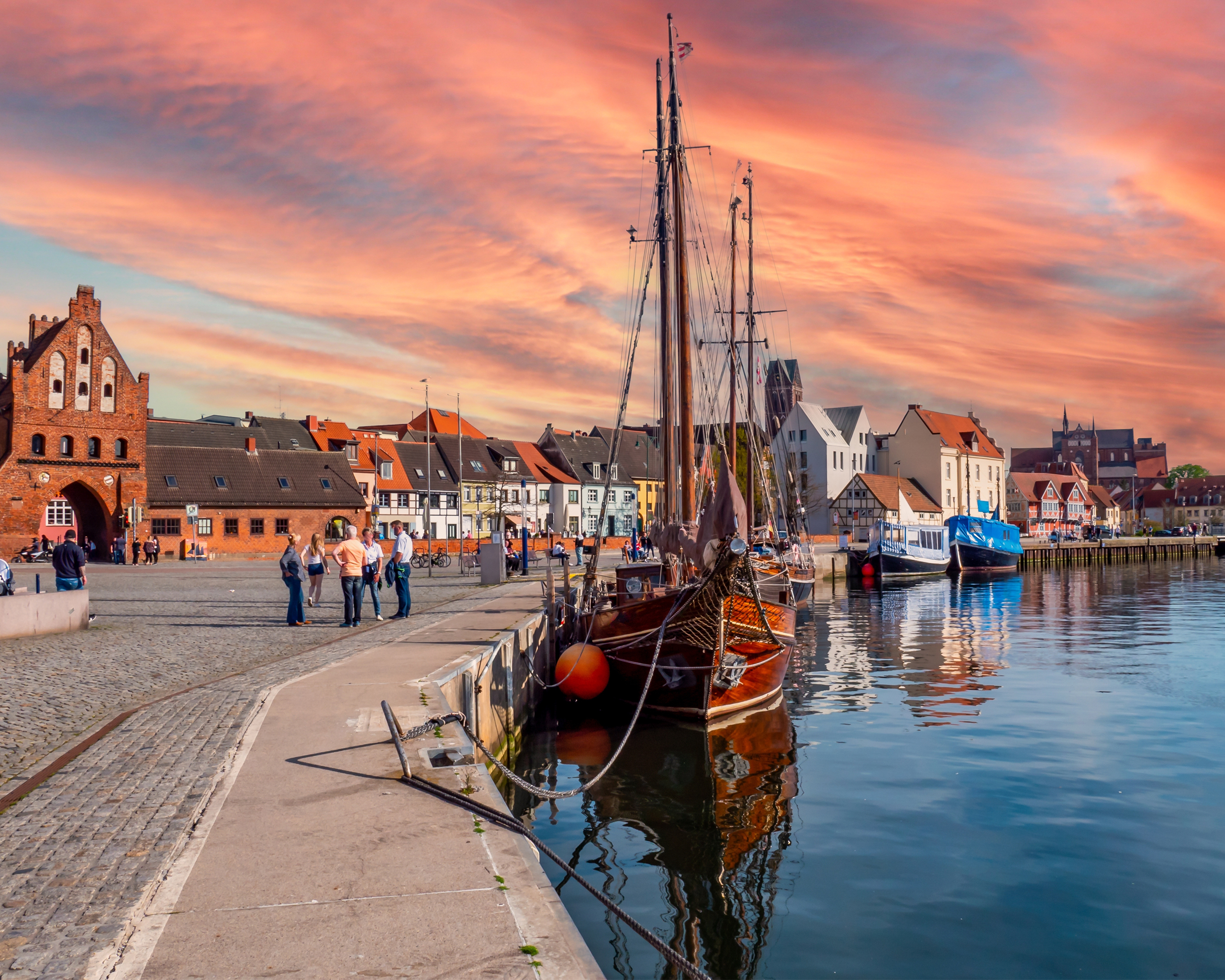 Historischer Hafen von Wismar mit traditionellen Segelschiffen, Backsteinhäusern und warmem Abendlicht am Wasser.