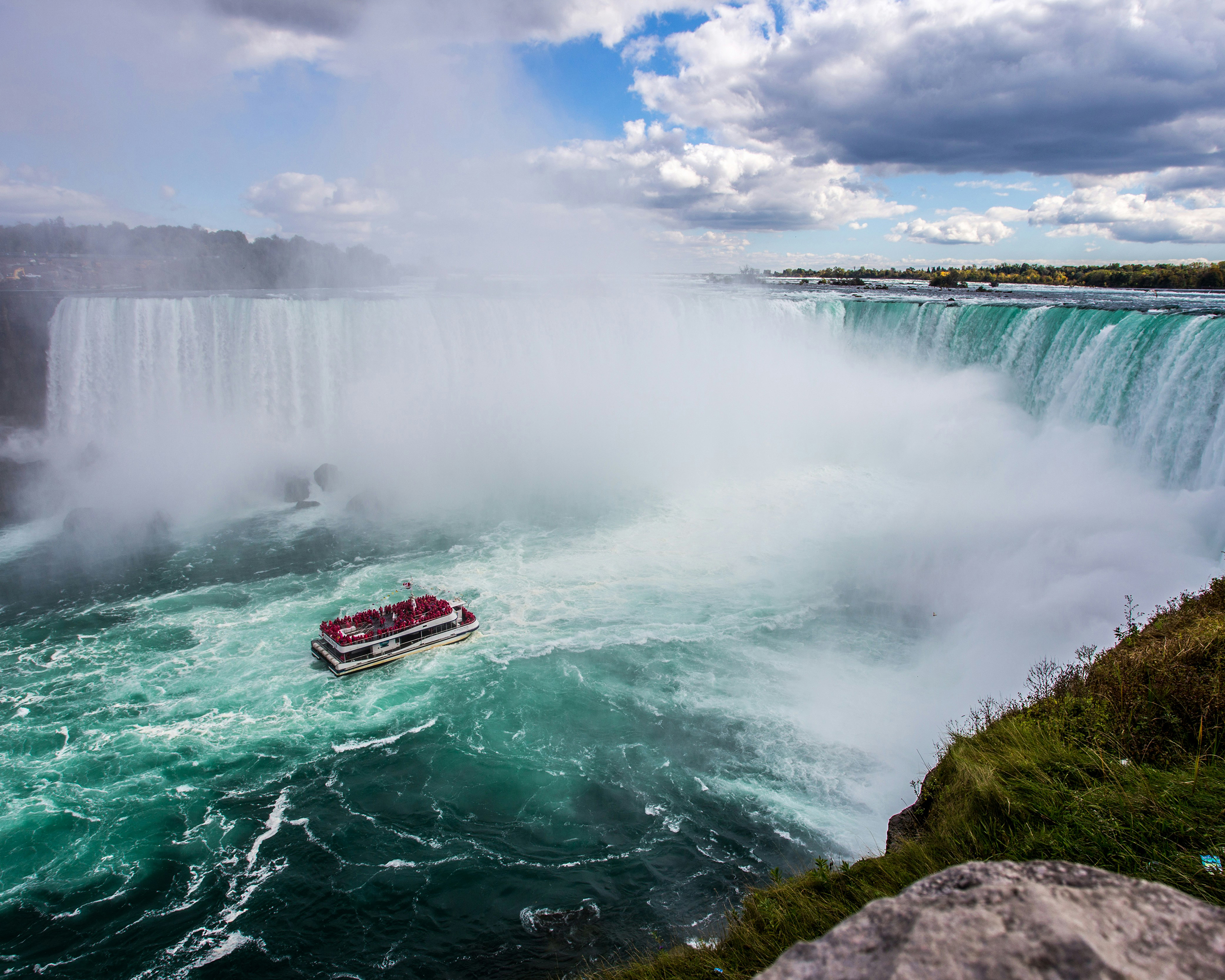Die mächtigen Niagarafälle mit aufsteigendem Sprühnebel, davor ein Ausflugsboot mit Besuchern auf dem türkisfarbenen Wasser.