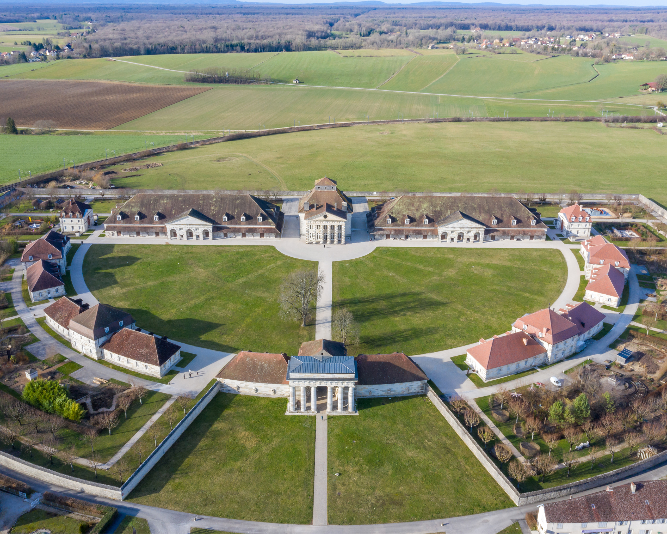 Luftaufnahme der halbkreisförmig angelegten Saline Royale d’Arc-et-Senans mit klassizistischen Gebäuden, grünen Rasenflächen und ländlicher Umgebung in Frankreich.