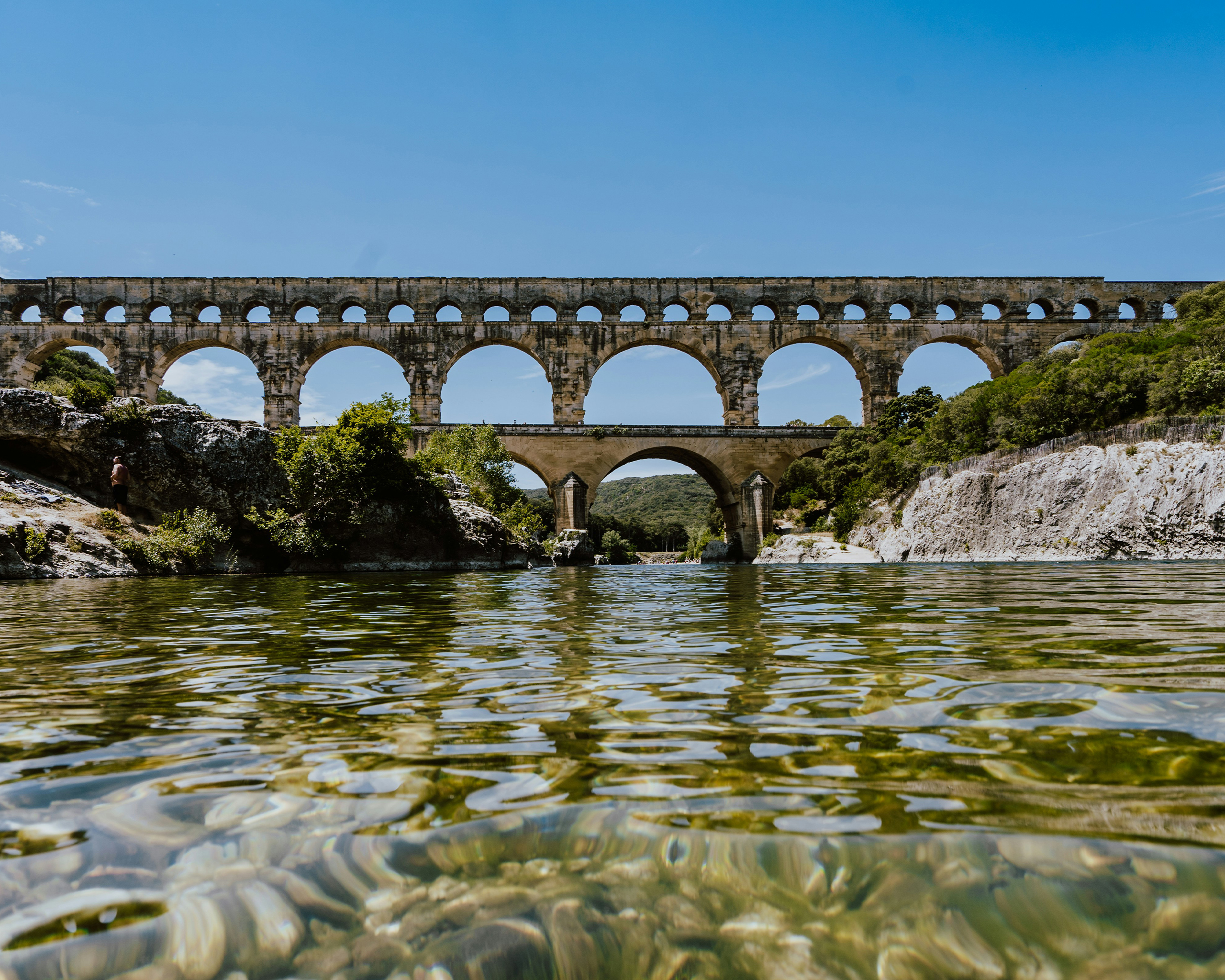 Römisches Aquädukt Pont du Gard mit imposanten Steinbögen über einem klaren Fluss, umgeben von Felsen und mediterraner Vegetation in der Provence.