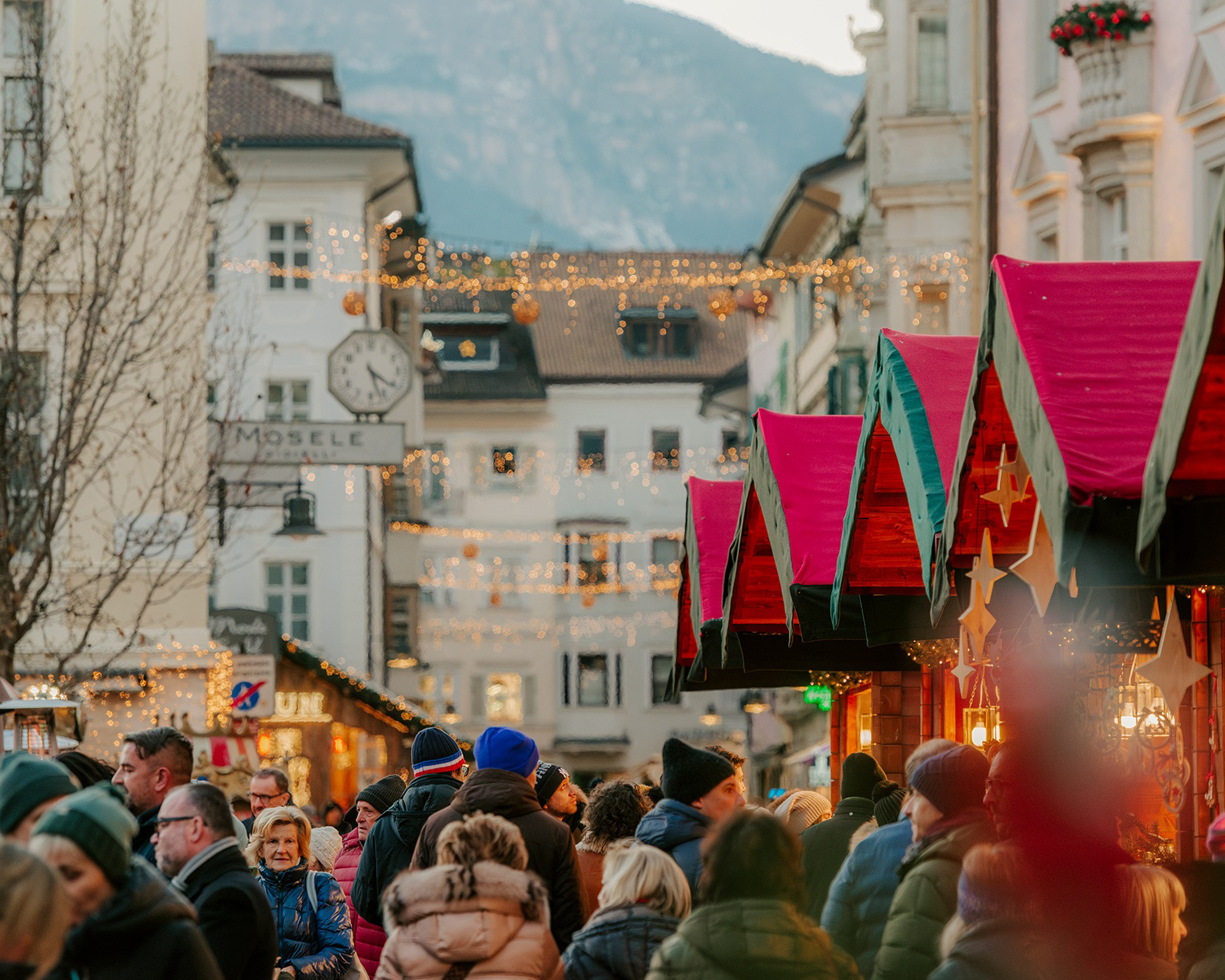 Belebter Weihnachtsmarkt in der Bozner Altstadt mit festlich geschmückten Marktständen, Lichterketten und winterlicher Bergkulisse im Hintergrund. © Mercatino di Natale Bolzano Bozen