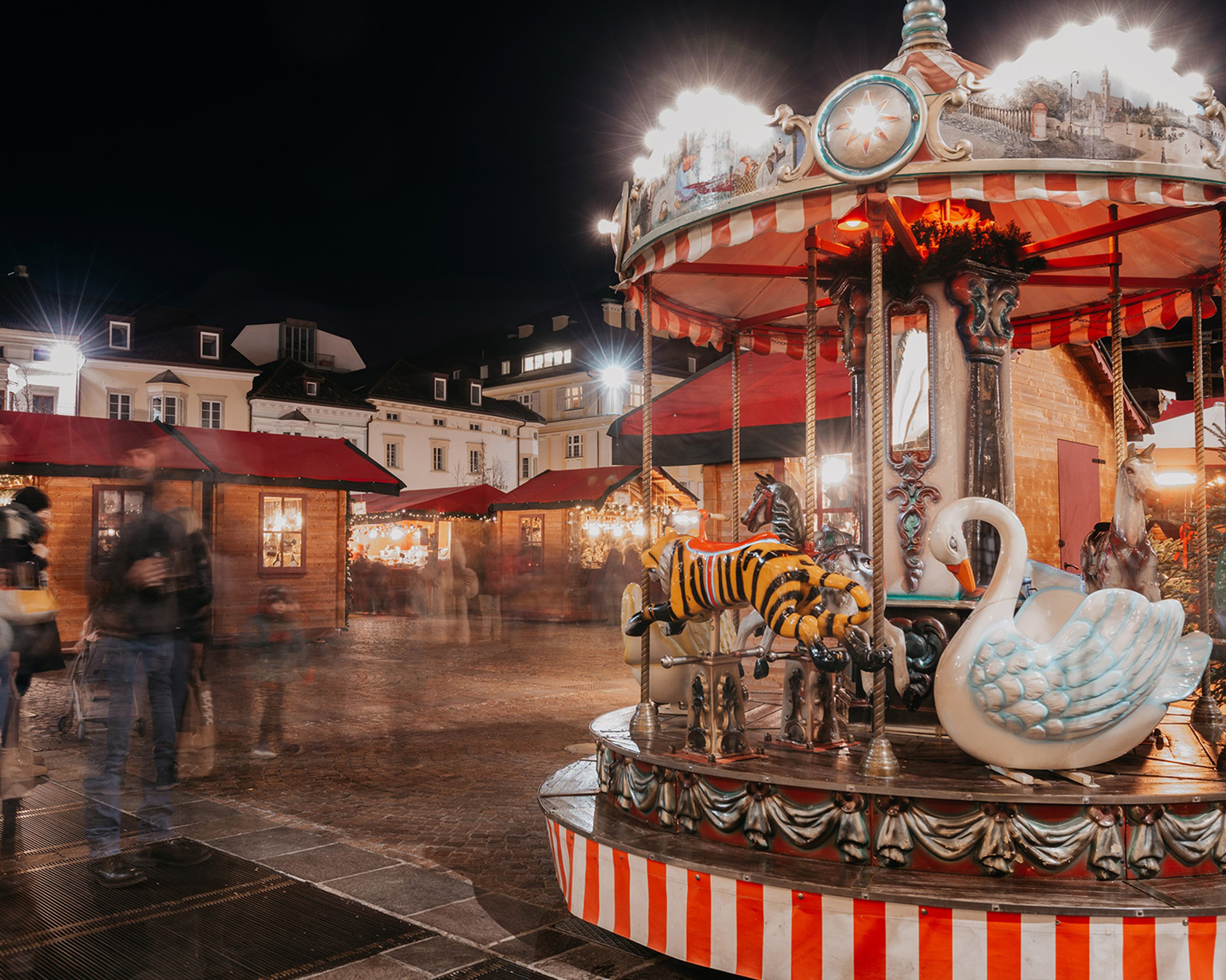 Historisches Kinderkarussell auf dem festlich beleuchteten Weihnachtsmarkt in Bozen bei Nacht, umgeben von weihnachtlich dekorierten Marktständen. © Mercatino di Natale Bolzano Bozen
