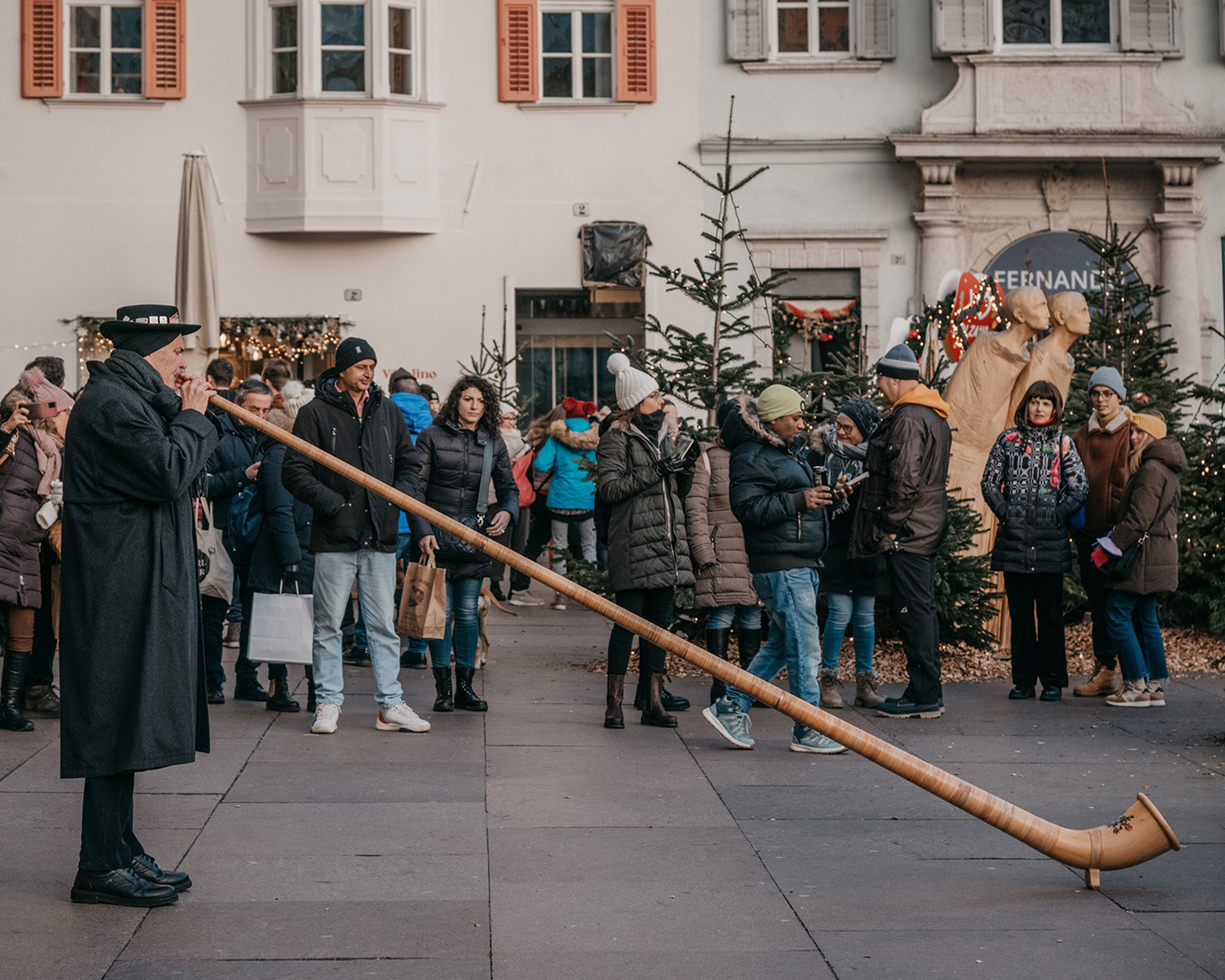 Musiker spielt ein Alphorn auf dem Weihnachtsmarkt in Bozen, während Besucher zwischen festlich geschmückten Tannenbäumen und Marktständen flanieren. © Mercatino di Natale Bolzano Bozen
