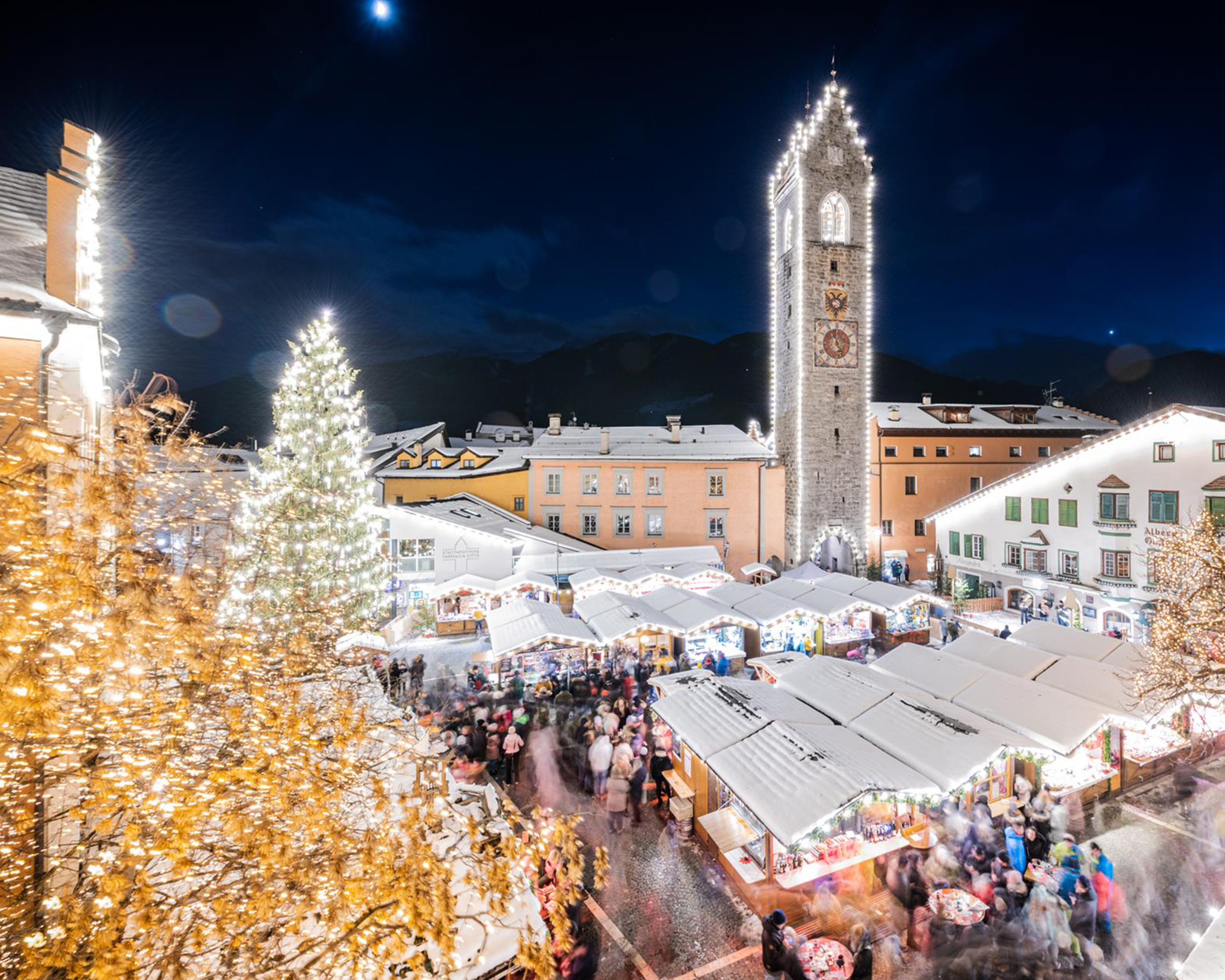 Festlich beleuchteter Weihnachtsmarkt auf dem Stadtplatz von Sterzing mit großem Christbaum, historischen Marktständen und dem Zwölferturm bei Nacht. © Wisthaler.com