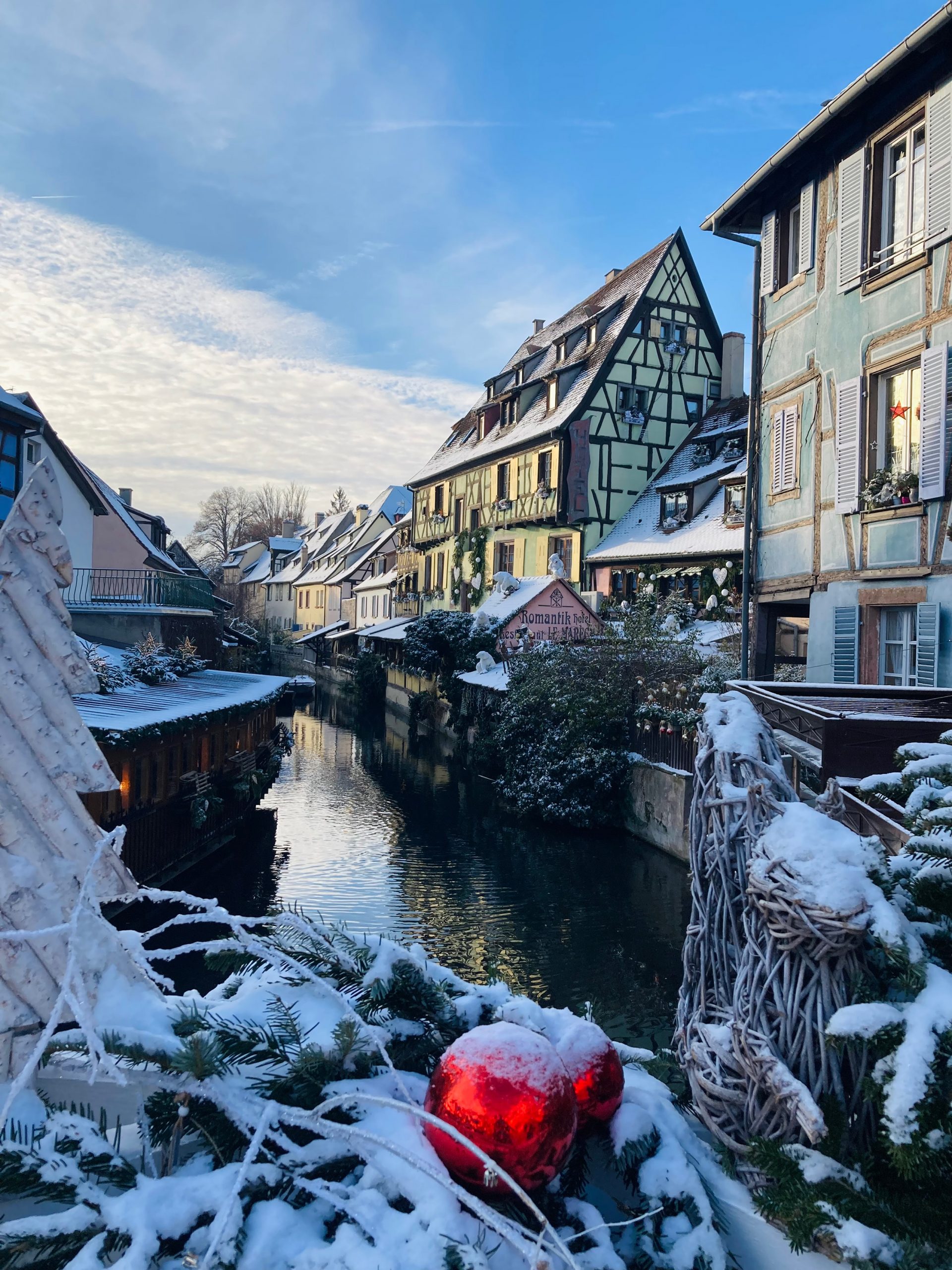 Verschneite Fachwerkhäuser spiegeln sich im Wasserkanal von Colmars Klein Venedig, festlich geschmückt mit roten Weihnachtskugeln