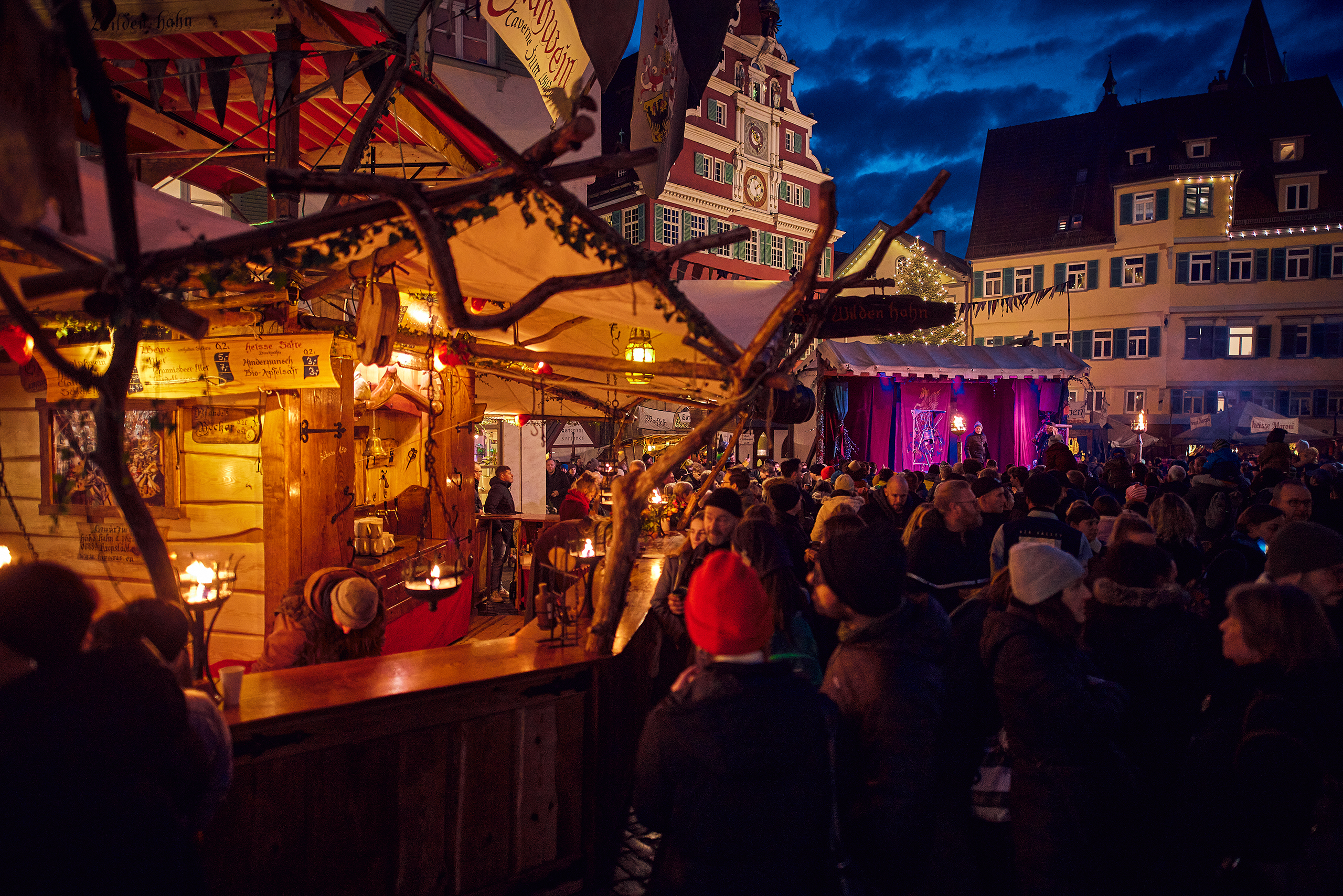 Stimmungsvolle Szene am Esslinger Mittelaltermarkt & Weihnachtsmarkt: Besucher genießen die Atmosphäre an einer mittelalterlich gestalteten Taverne, während auf der Bühne vor dem Alten Rathaus ein Abendprogramm stattfindet.
