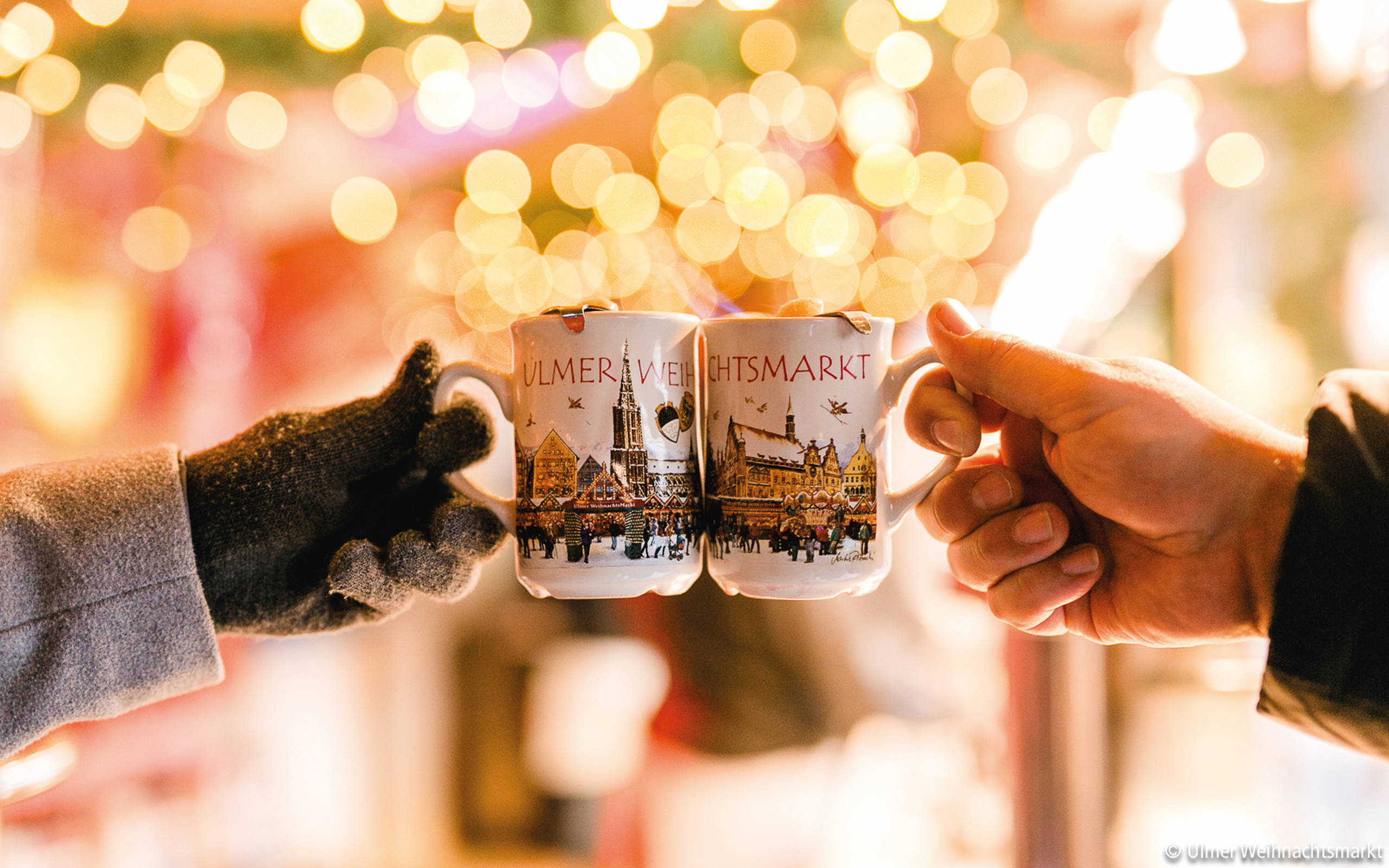Zwei Tassen Glühwein mit Ulmer Weihnachtsmarkt-Motiv stoßen an – Genuss und Geselligkeit in festlicher Stimmung
