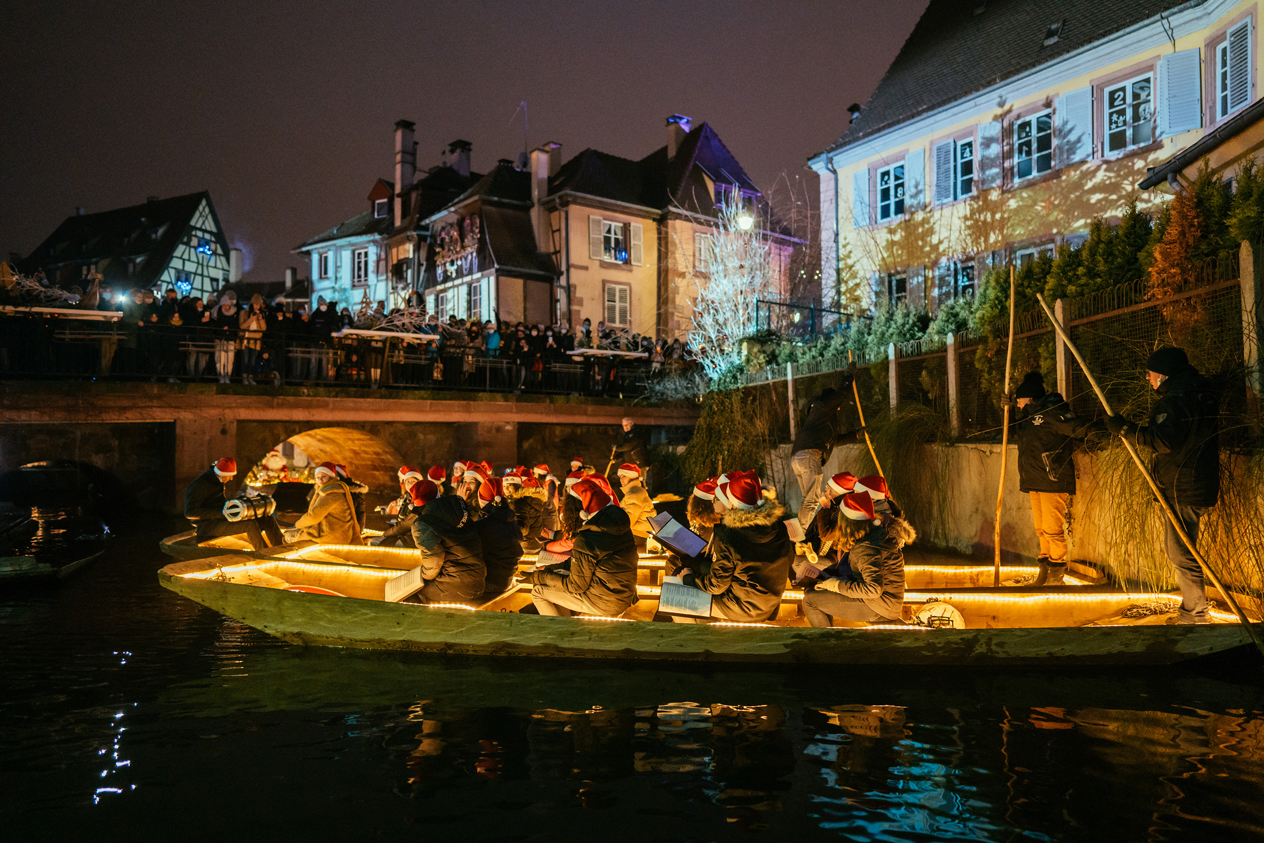 Stimmungsvoll beleuchtete Boote mit Weihnachtschören ziehen durch Colmars Altstadt, während Besucher das Spektakel vom Ufer aus bestaunen