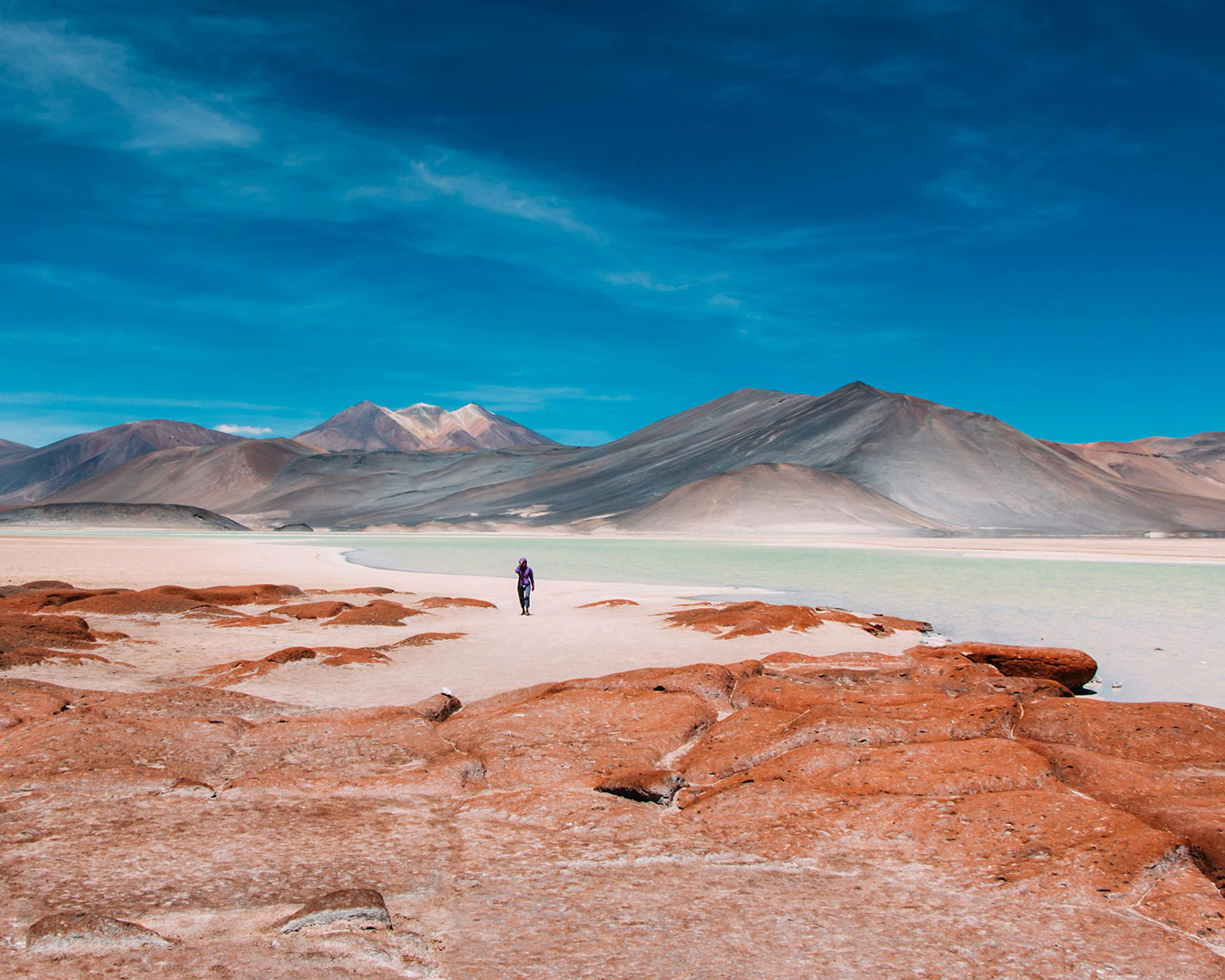 Farbenprächtige Wüstenlandschaft der Atacama in Chile mit türkisfarbenem Salzsee, rötlichen Felsen und imposanten Andenbergen.