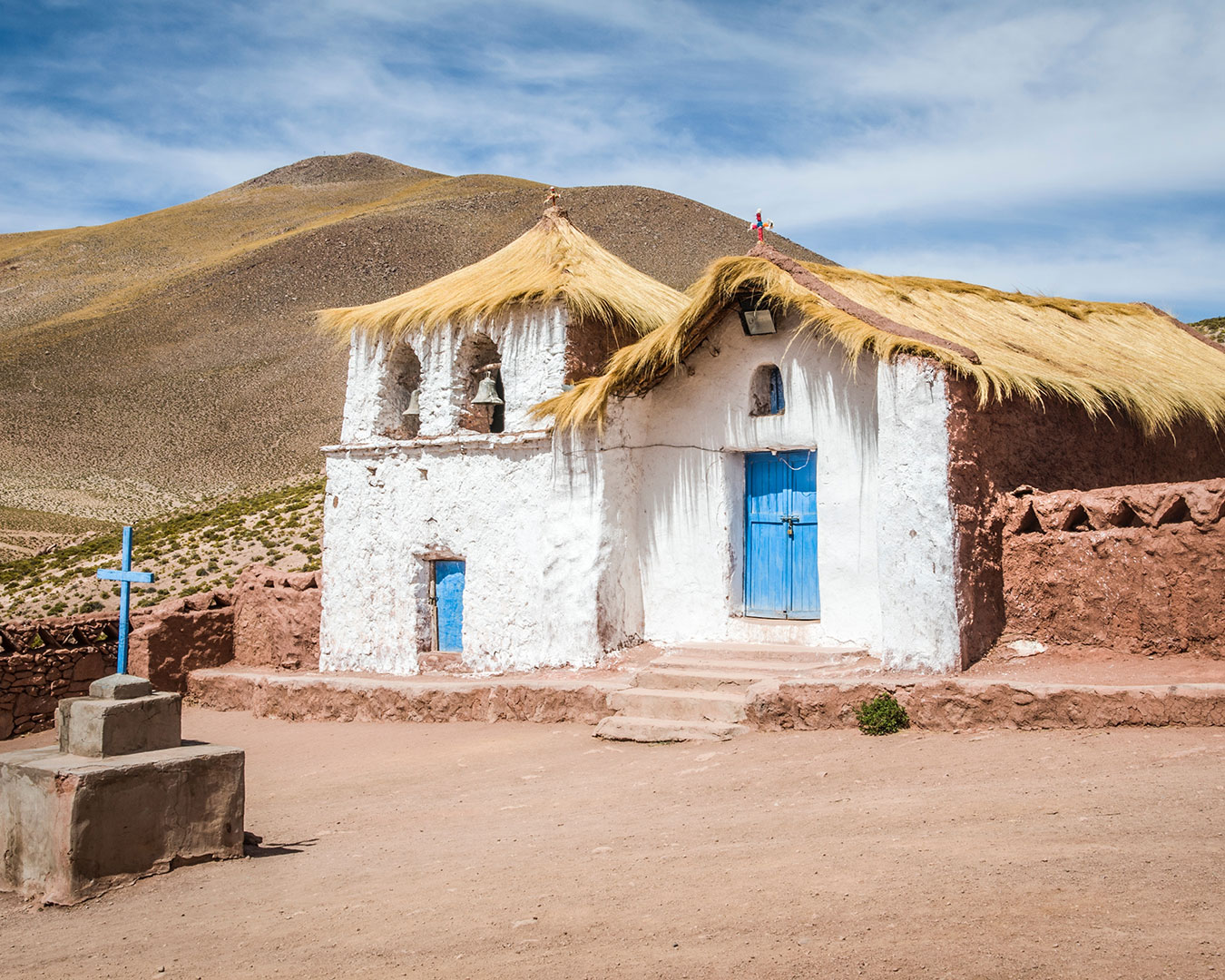Traditionelle weiß verputzte Kirche von Machuca in Chile mit Strohdach, blauer Holztür und Glockenturm vor karger Andenlandschaft.