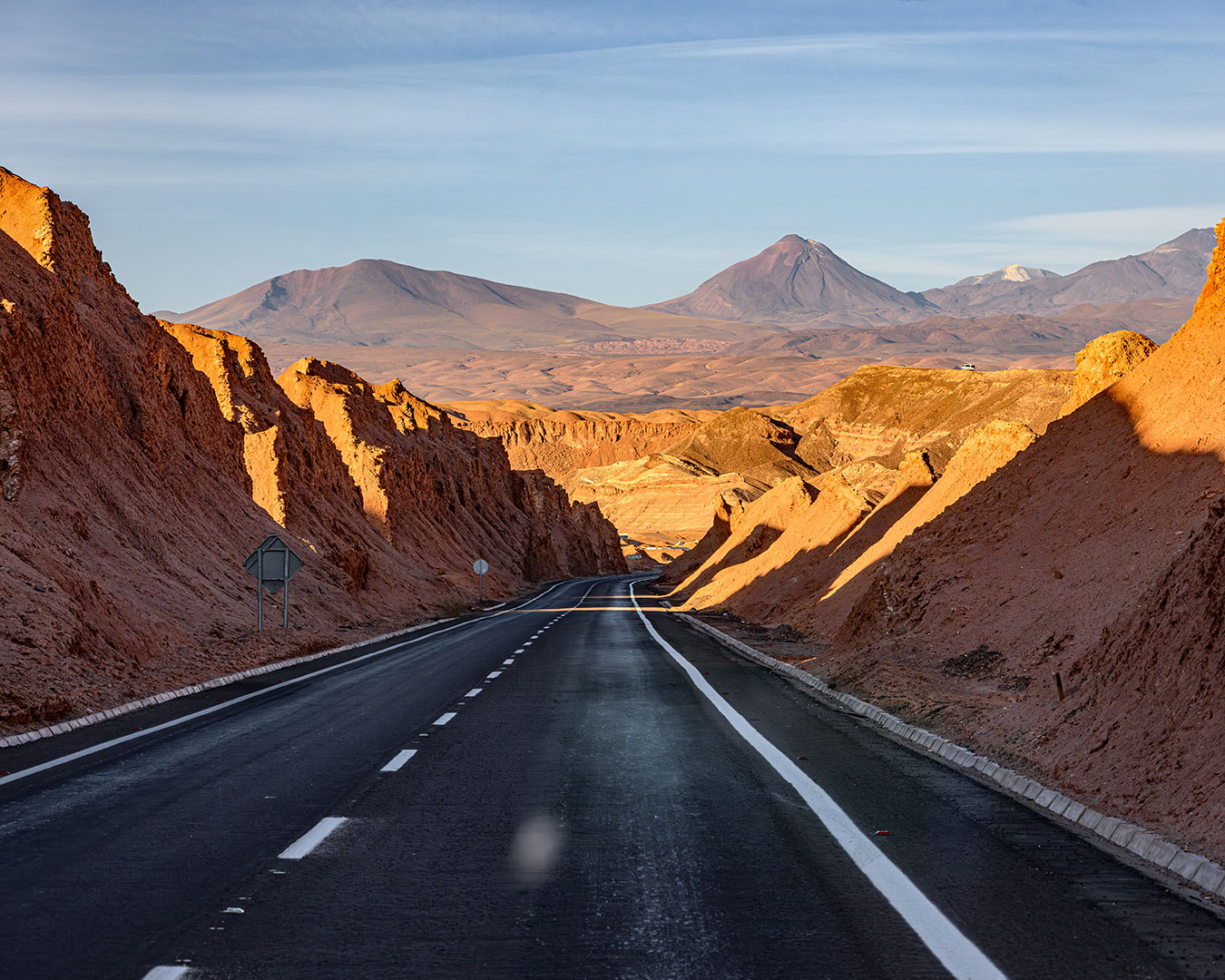 Asphaltierte Straße durch die rötliche Atacama-Wüste zwischen Calama und San Pedro de Atacama mit Vulkanbergen im Hintergrund.