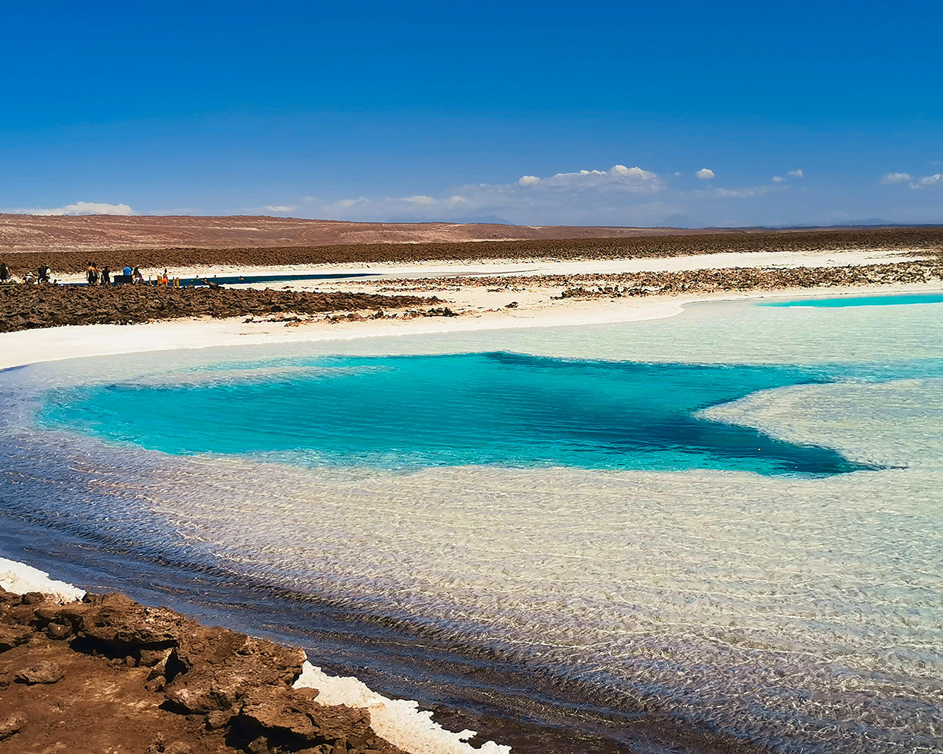 Türkisblaue Salzlagune mit weißen Salzablagerungen und dunklen Felsen in der Atacama-Wüste in Chile.