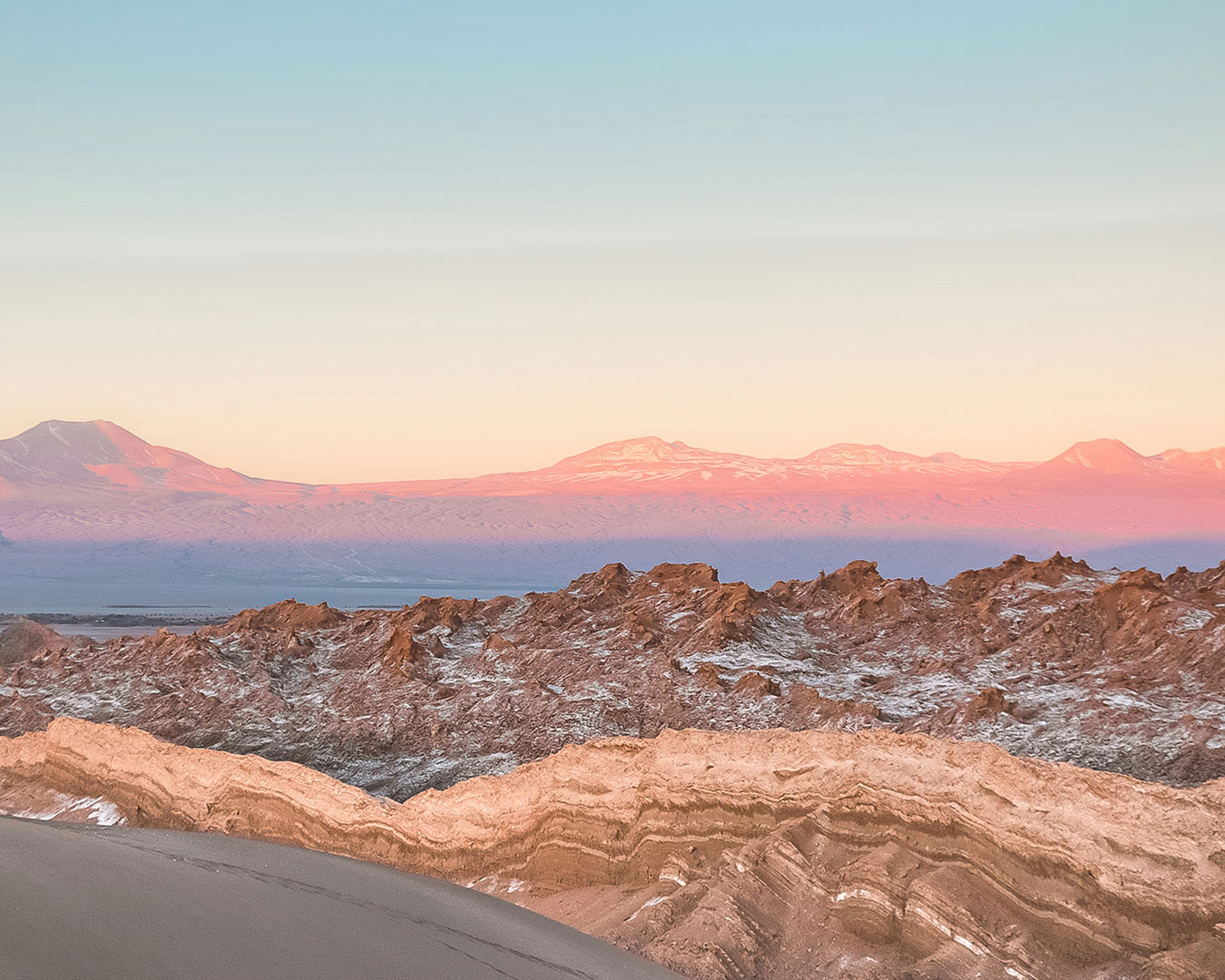 Zerklüftete Felsformationen im Valle de la Luna in der Atacama-Wüste mit rosafarben beleuchteten Andenbergen.