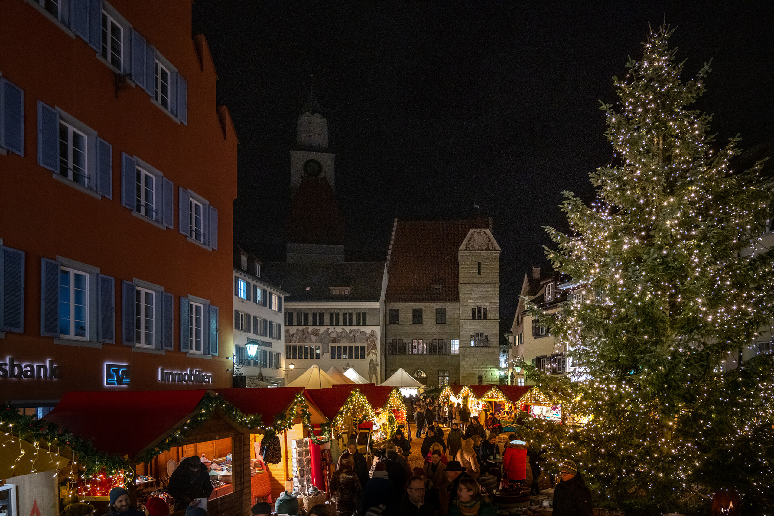 Romantische Abendstimmung auf dem Überlinger Weihnachtsmarkt: Menschen flanieren zwischen hell erleuchteten Ständen, im Hintergrund funkelt der große Weihnachtsbaum